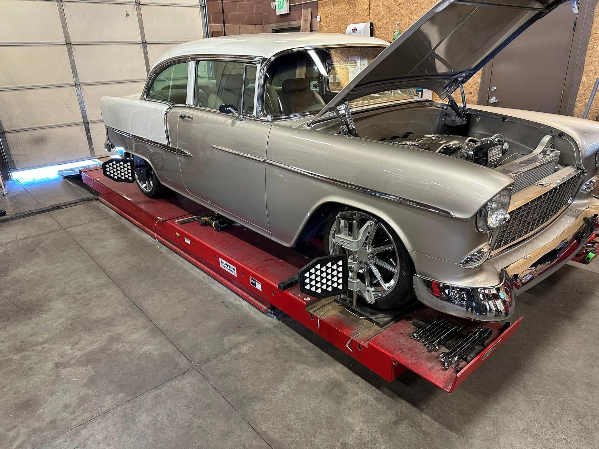 Silver classic car on a lift; hood open, wheels aligned, in a garage.