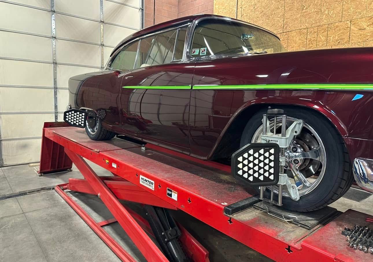Burgundy classic car on a red lift undergoing wheel alignment in a garage.