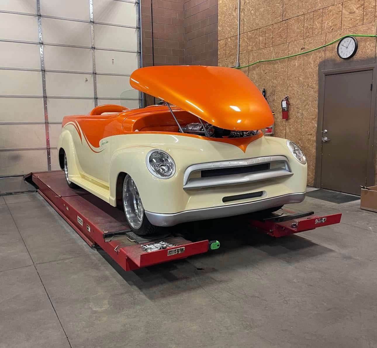 Cream and orange custom car with hood up on a red lift inside a garage.