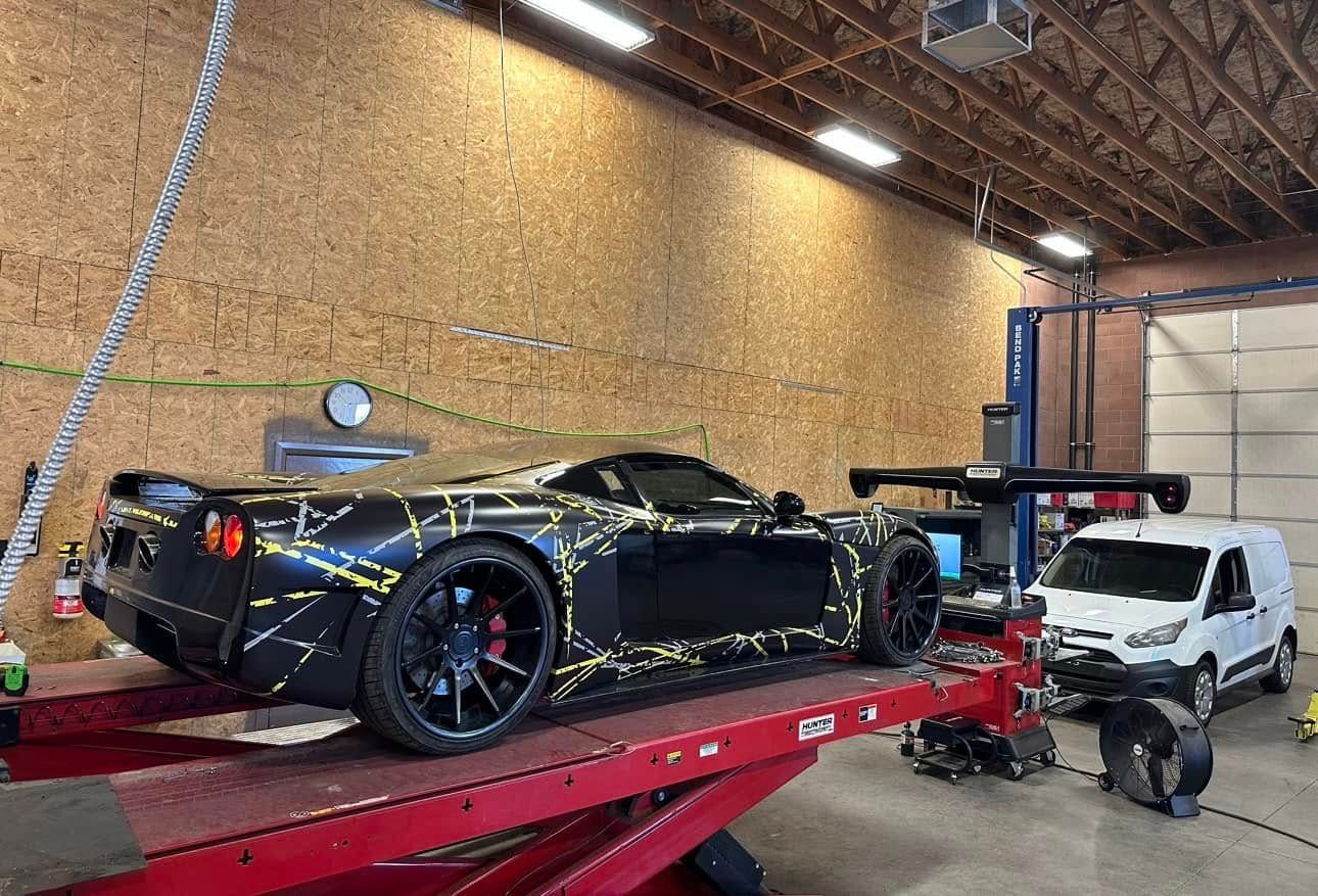 Black sports car with gold accents on a lift in a garage. A white van is parked nearby.