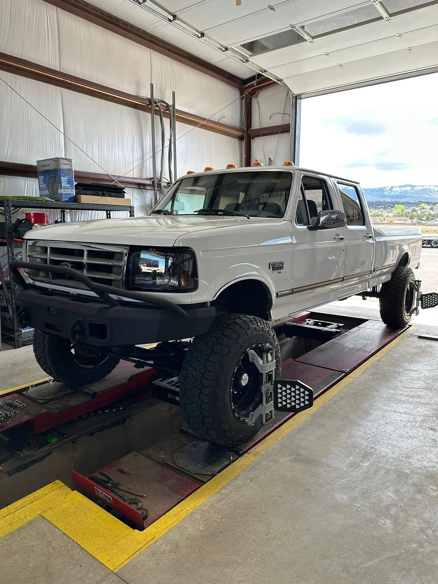 White lifted Ford truck inside a garage for alignment, large tires, black front bumper, and open bay door.