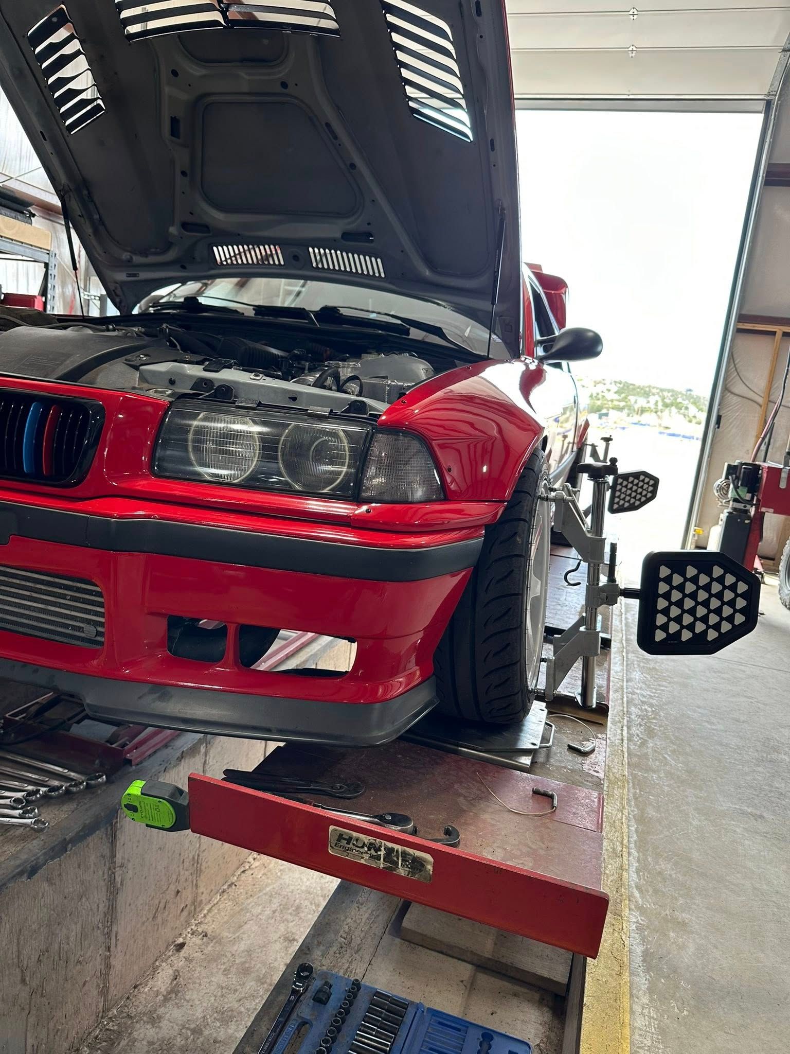 Red BMW E36 on a car lift for wheel alignment, in a garage. Hood is open.