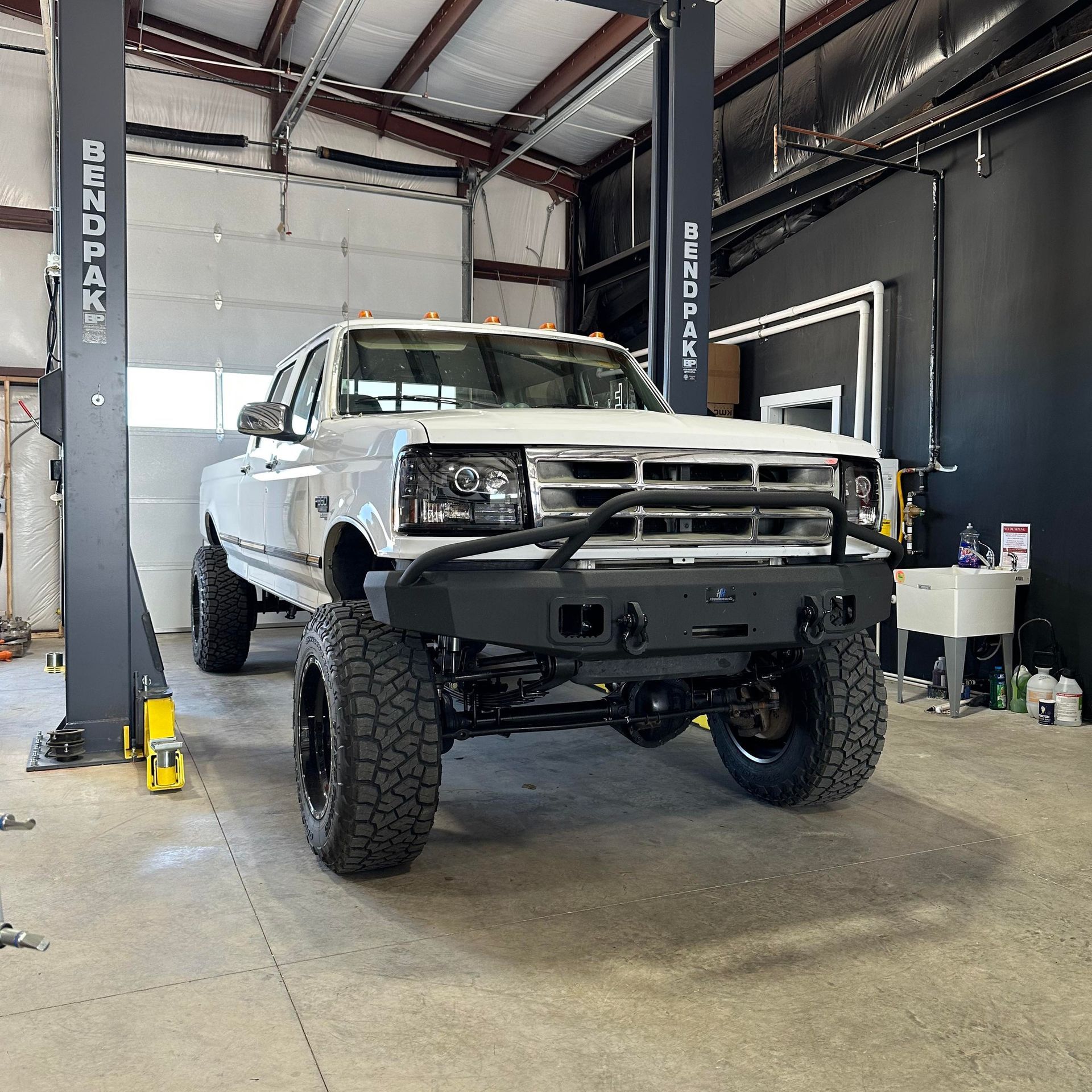 White pickup truck on a lift in a garage. Black bumper, tires, lift posts, and details.