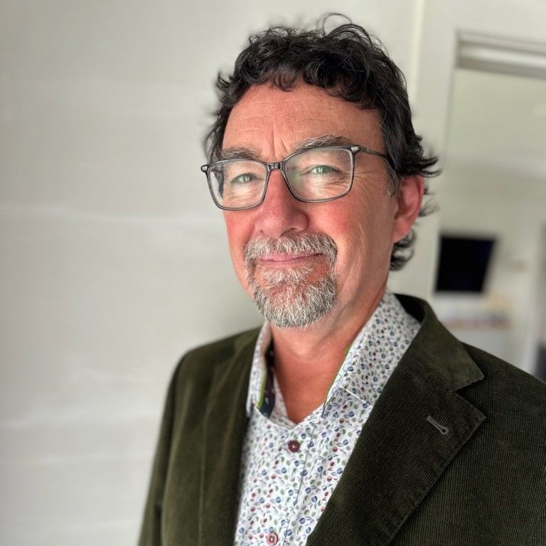 Man in glasses, green jacket, patterned shirt, slight smile, indoors.