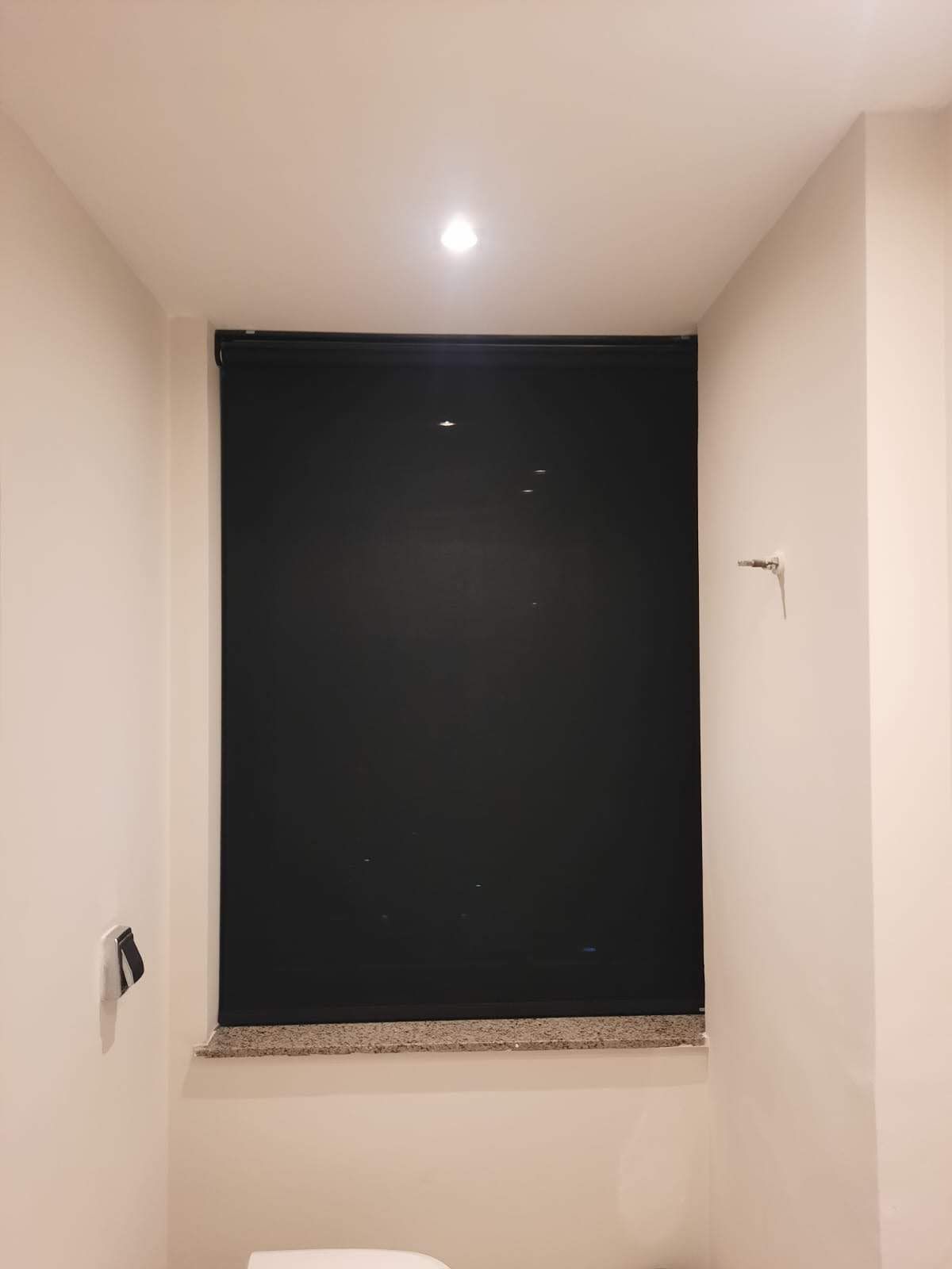 Uma janela com uma persiana preta fechada. Paredes bege e uma pequena luminária são visíveis.
