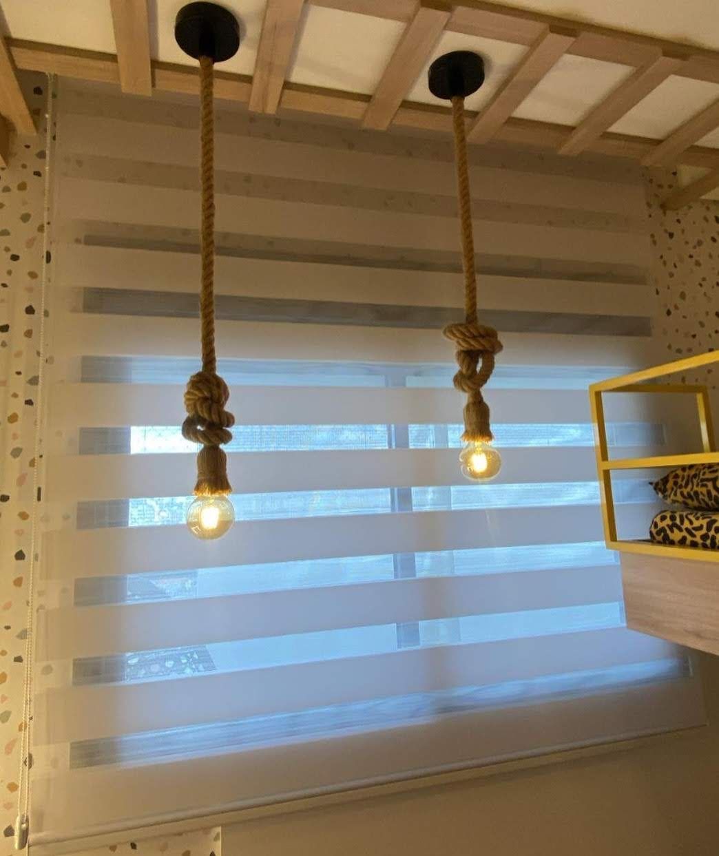 Duas luminárias pendentes de corda estão penduradas em frente a uma persiana listrada. As lâmpadas estão iluminadas e o ambiente transmite uma sensação aconchegante.