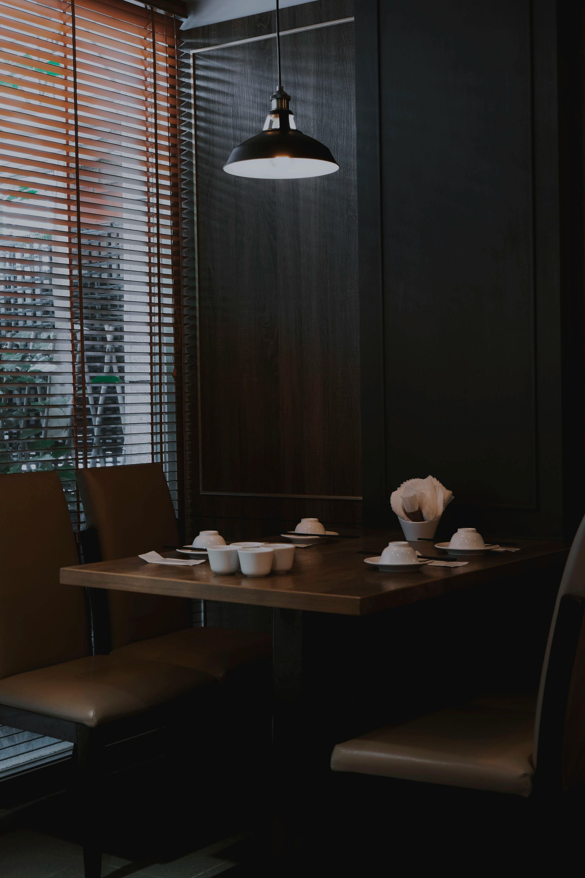 Ambiente escuro de restaurante. Uma mesa quadrada de madeira está posta com pratos, sob uma luminária pendente. Cadeiras ladeiam a mesa.