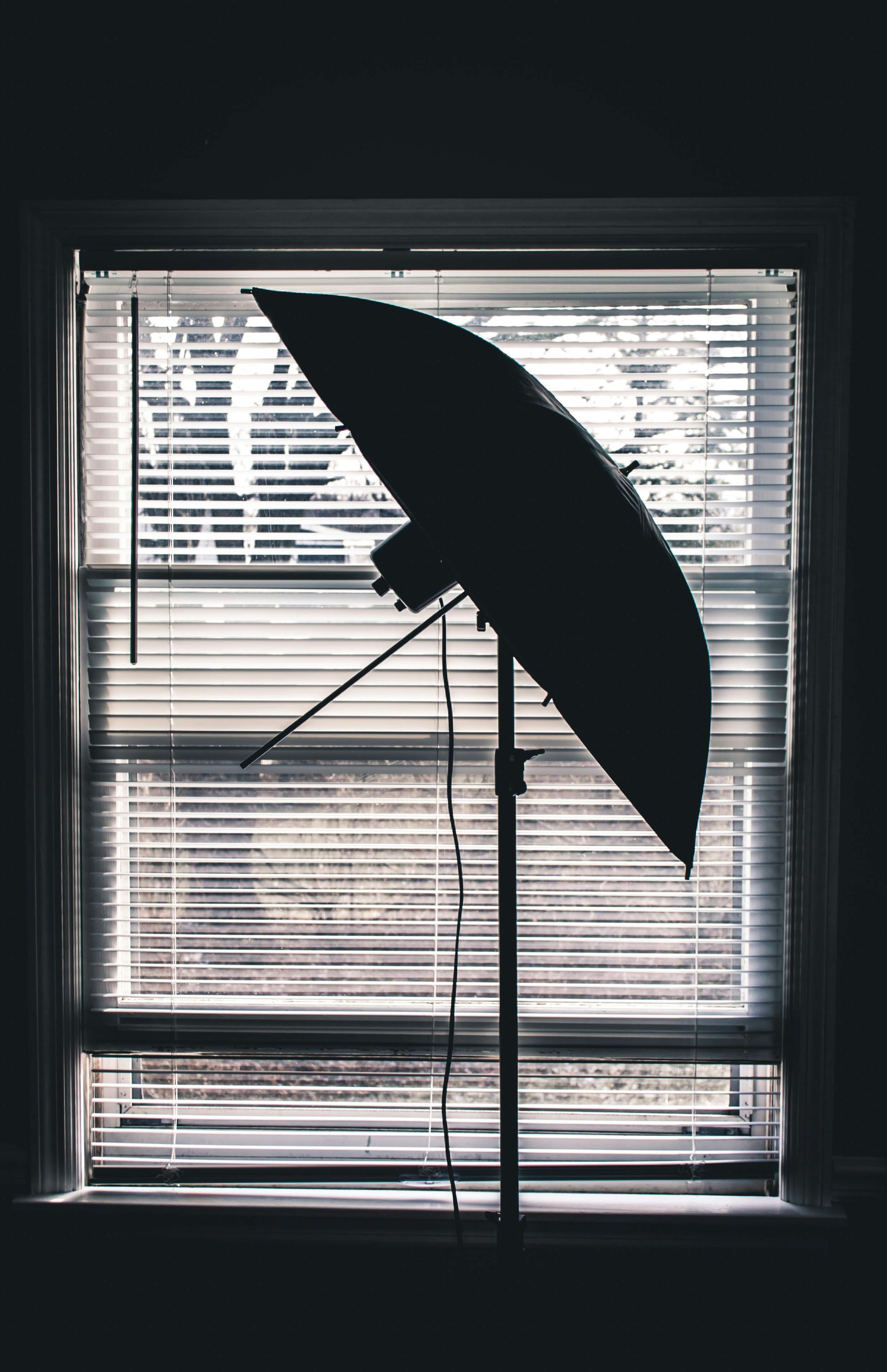 Silhueta de um guarda-chuva fotográfico em um suporte em frente a uma janela com persianas.