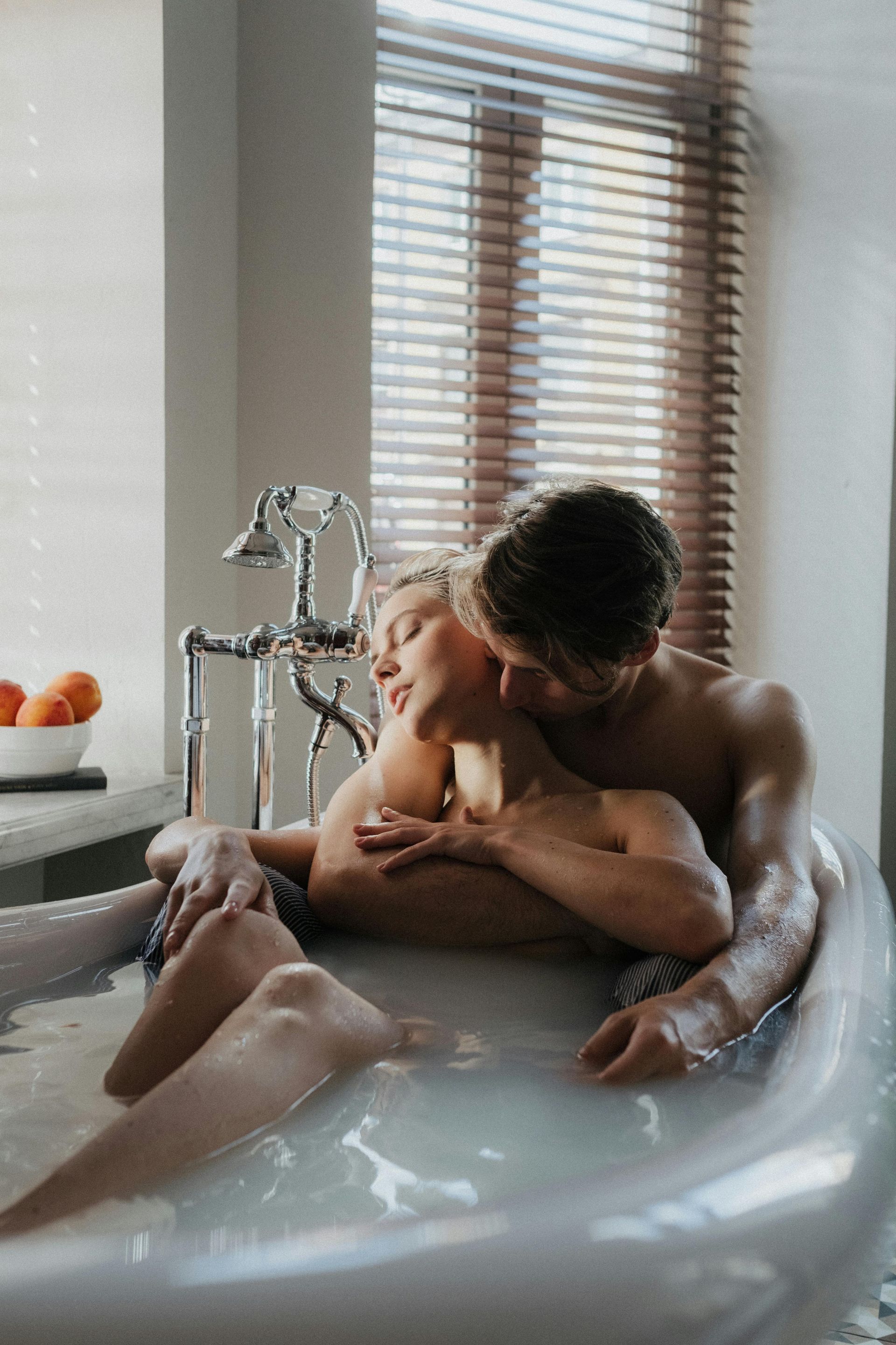 Casal se abraça intimamente em uma banheira. O homem beija o pescoço da mulher. Uma luz suave preenche o banheiro.