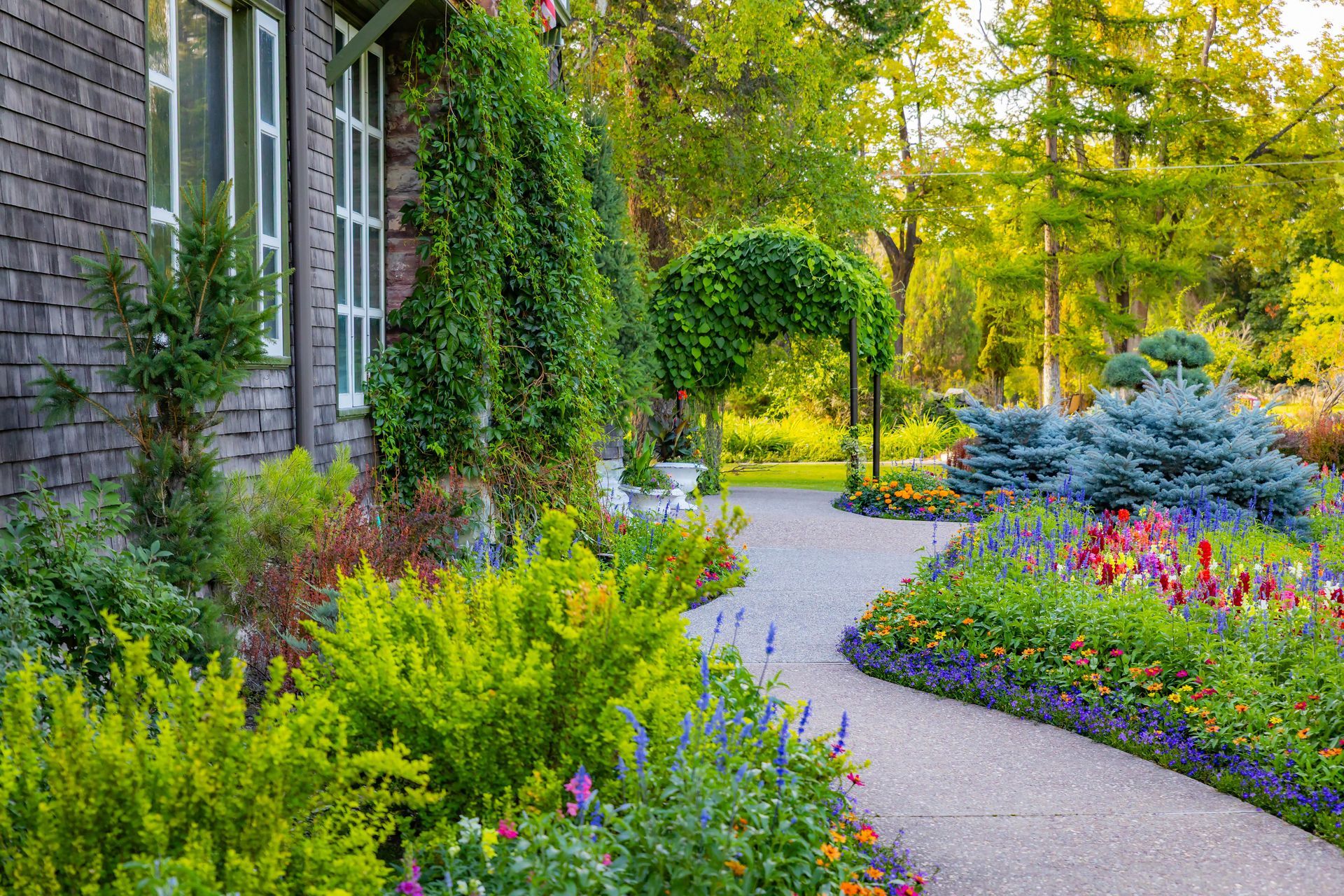 Stone walkway winds through a colorful garden beside a wood-shingled house.