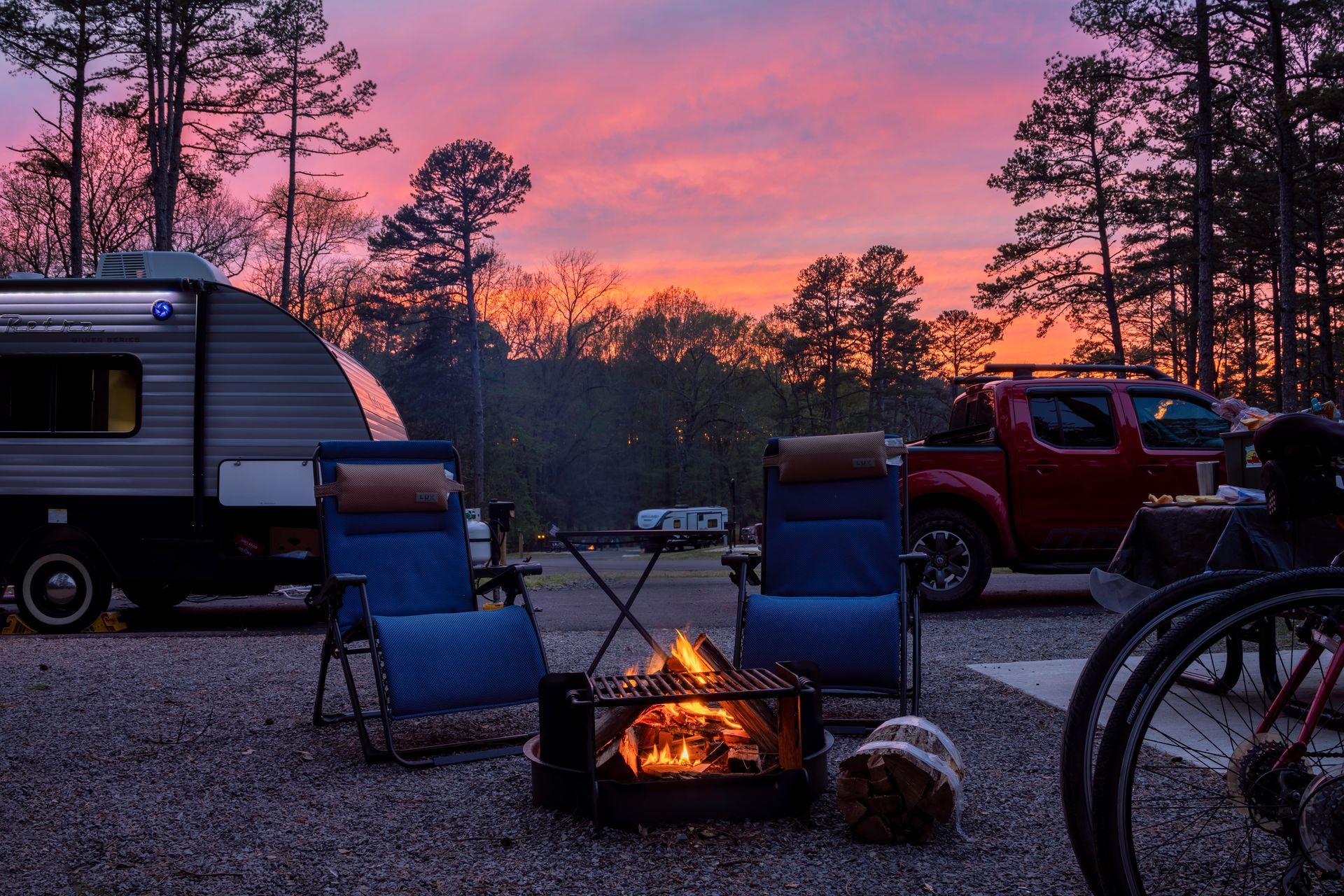 Camping scene at dusk: campfire, chairs, camper, red truck, bicycles, and pink and orange sky.