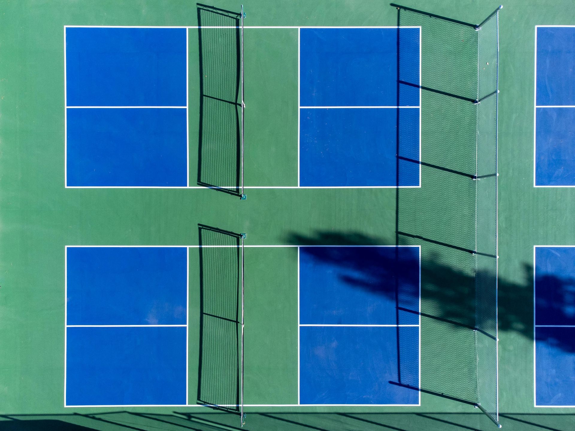 Overhead view of several blue pickleball courts with nets on a green surface, casting shadows.