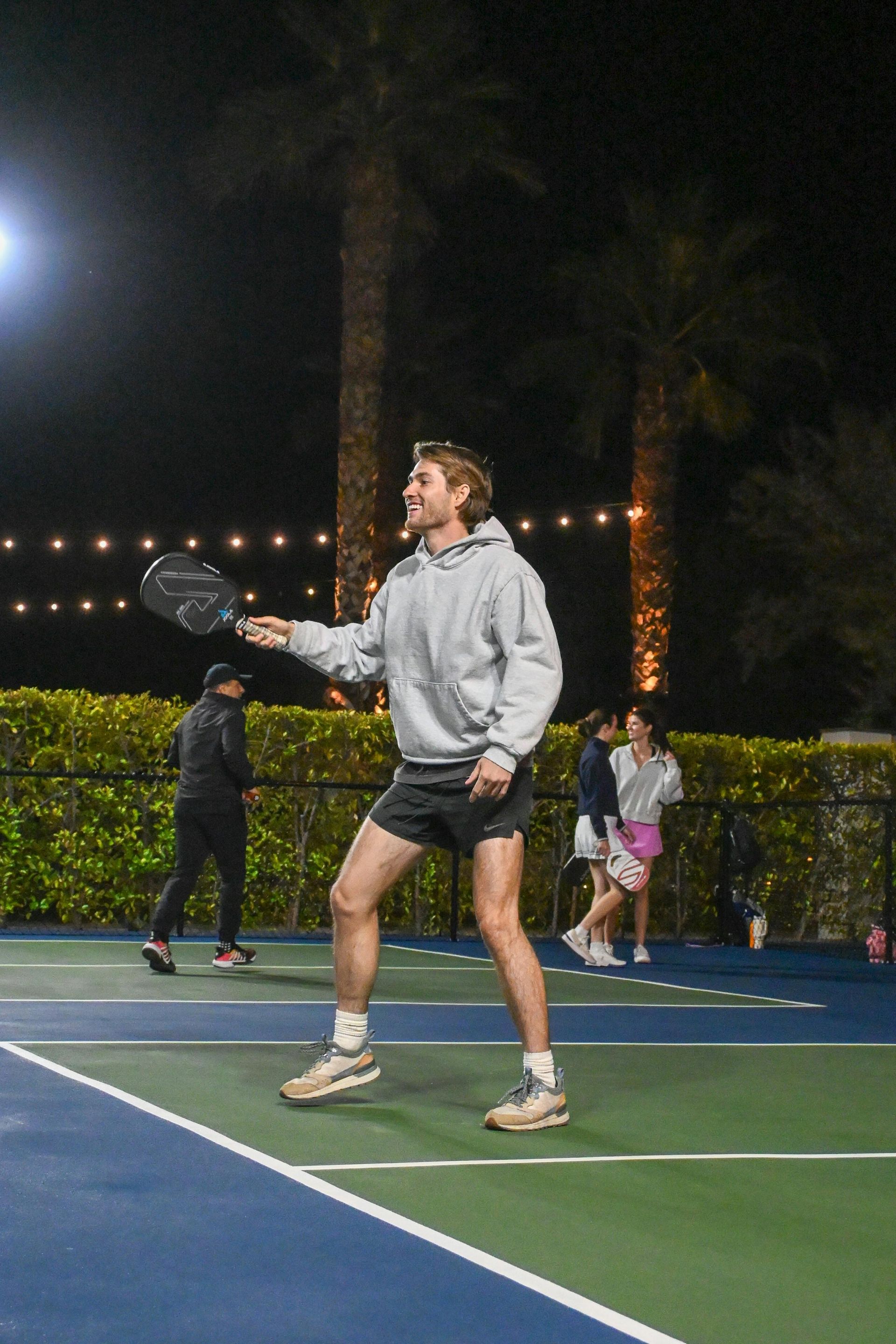 Man playing pickleball outdoors at night, holding paddle. Smiling, wearing grey hoodie and black shorts.
