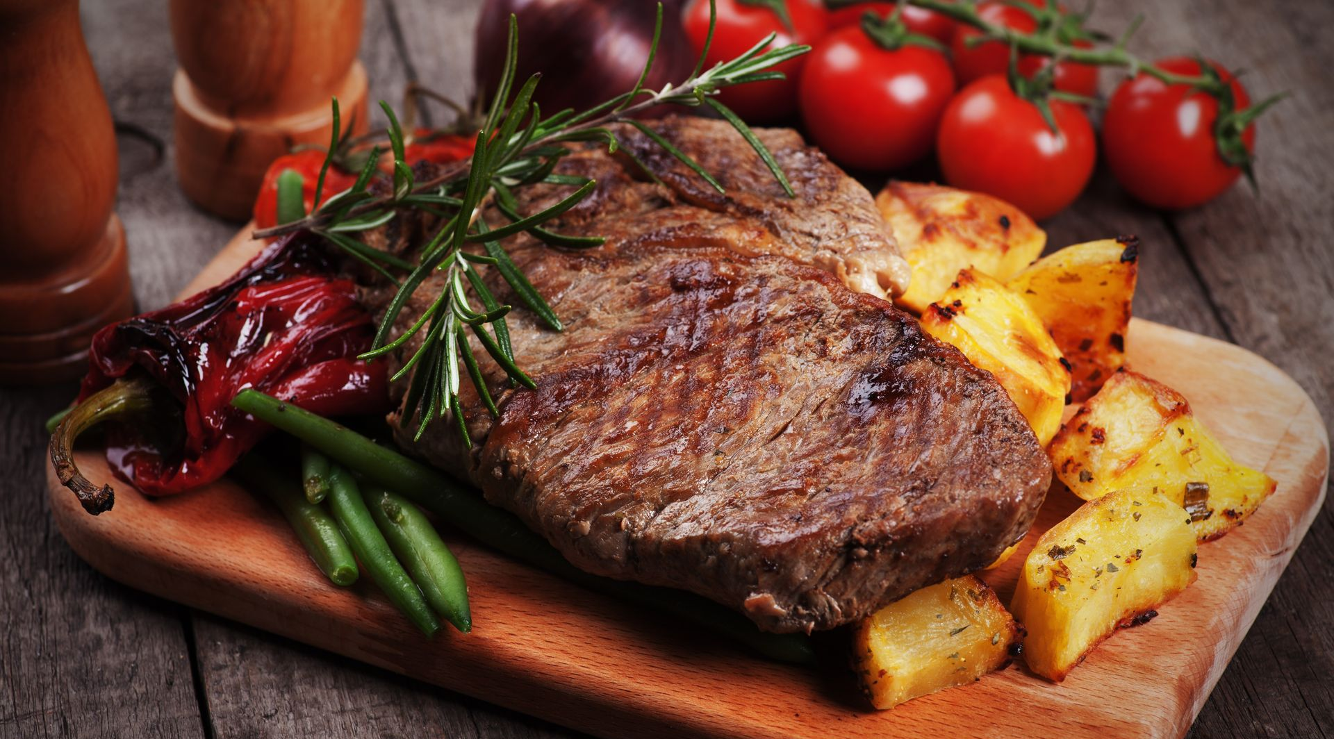 Steak dinner: grilled steak, roasted potatoes, tomatoes, rosemary, red peppers, green beans on a wooden board.