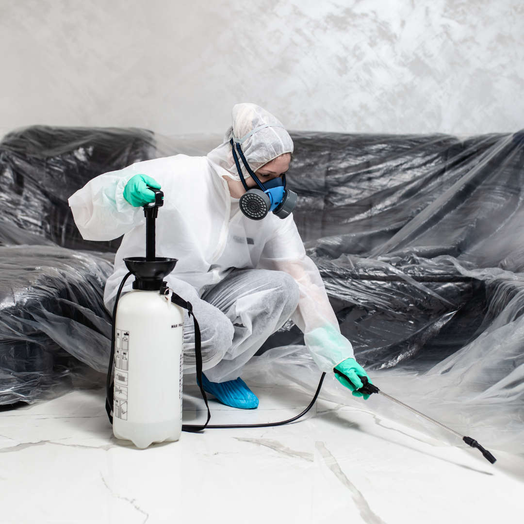 A man in a protective suit is spraying a couch with a sprayer.