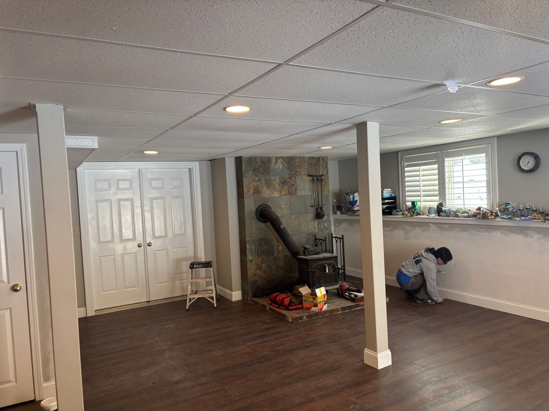 A person kneels to paint a basement wall near a wood-burning stove set against a stone fireplace.