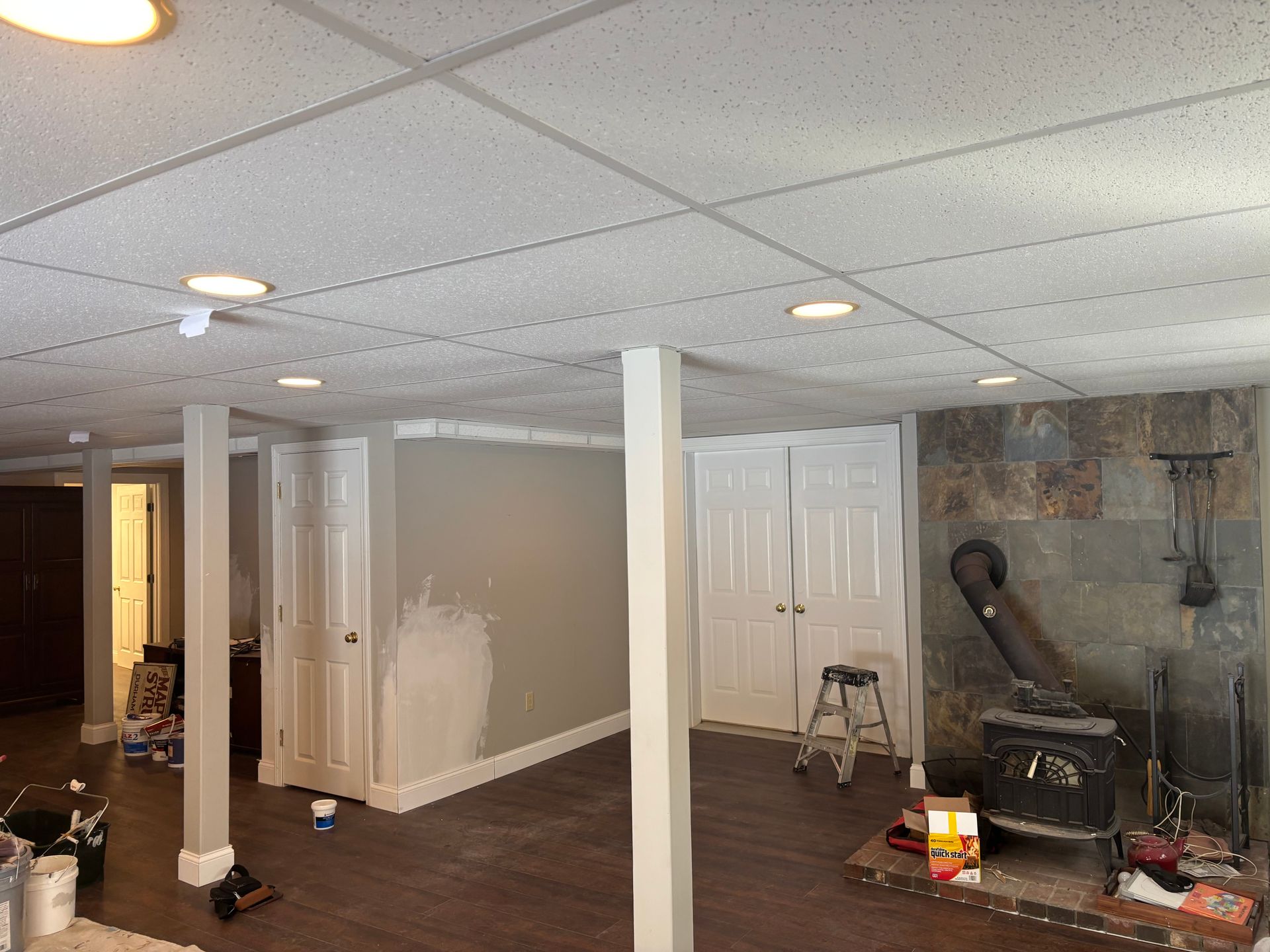 A basement renovation in progress with white panel ceilings, gray walls, support pillars, a wood stove, and closed doors.