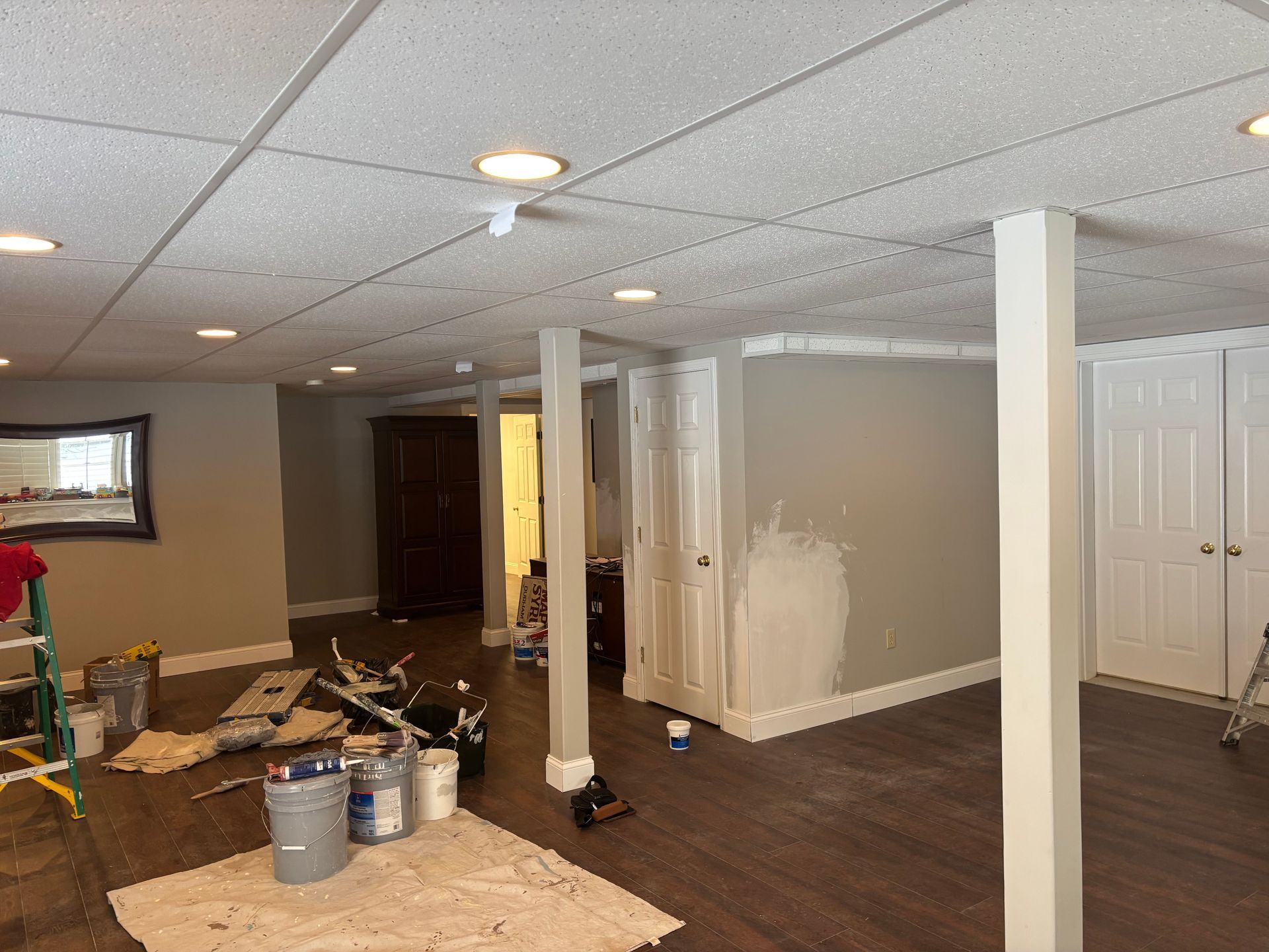 A basement room under renovation with beige walls, white support columns, a drop ceiling, and painting supplies on the floor.