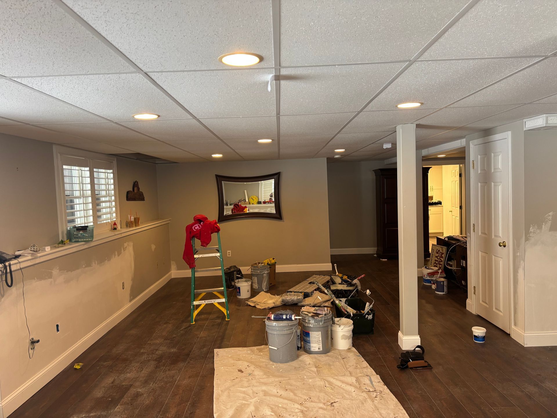 A basement renovation with dark wood floors, gray walls, a drop ceiling, a ladder, paint supplies, and an open doorway.