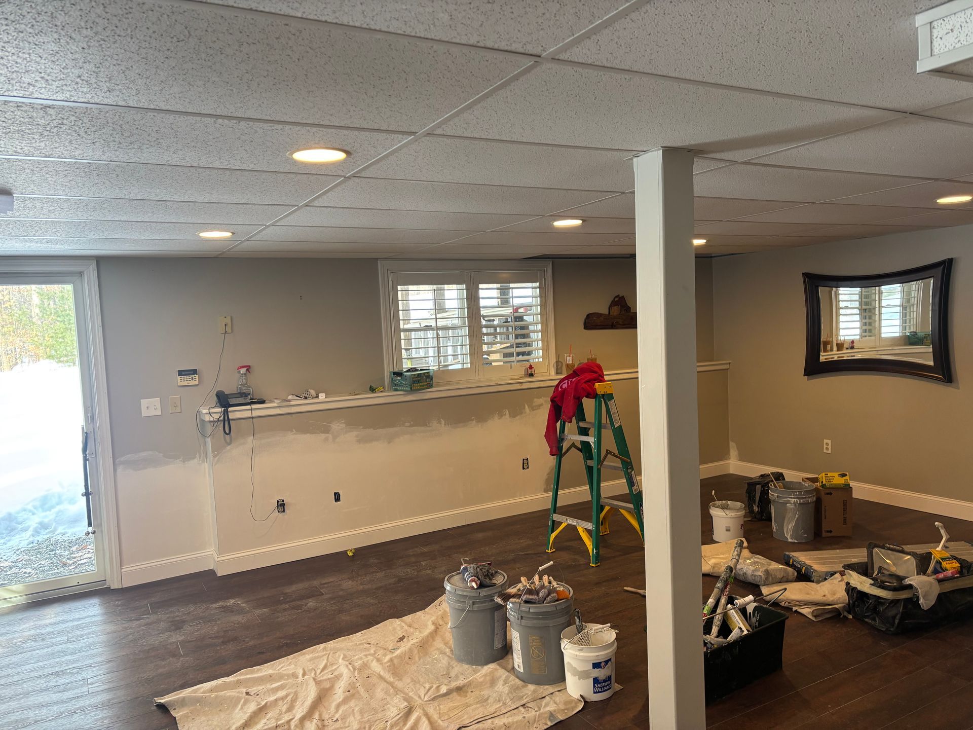 A basement under renovation with a bar counter, gray walls, a step ladder, and painting supplies on the floor.