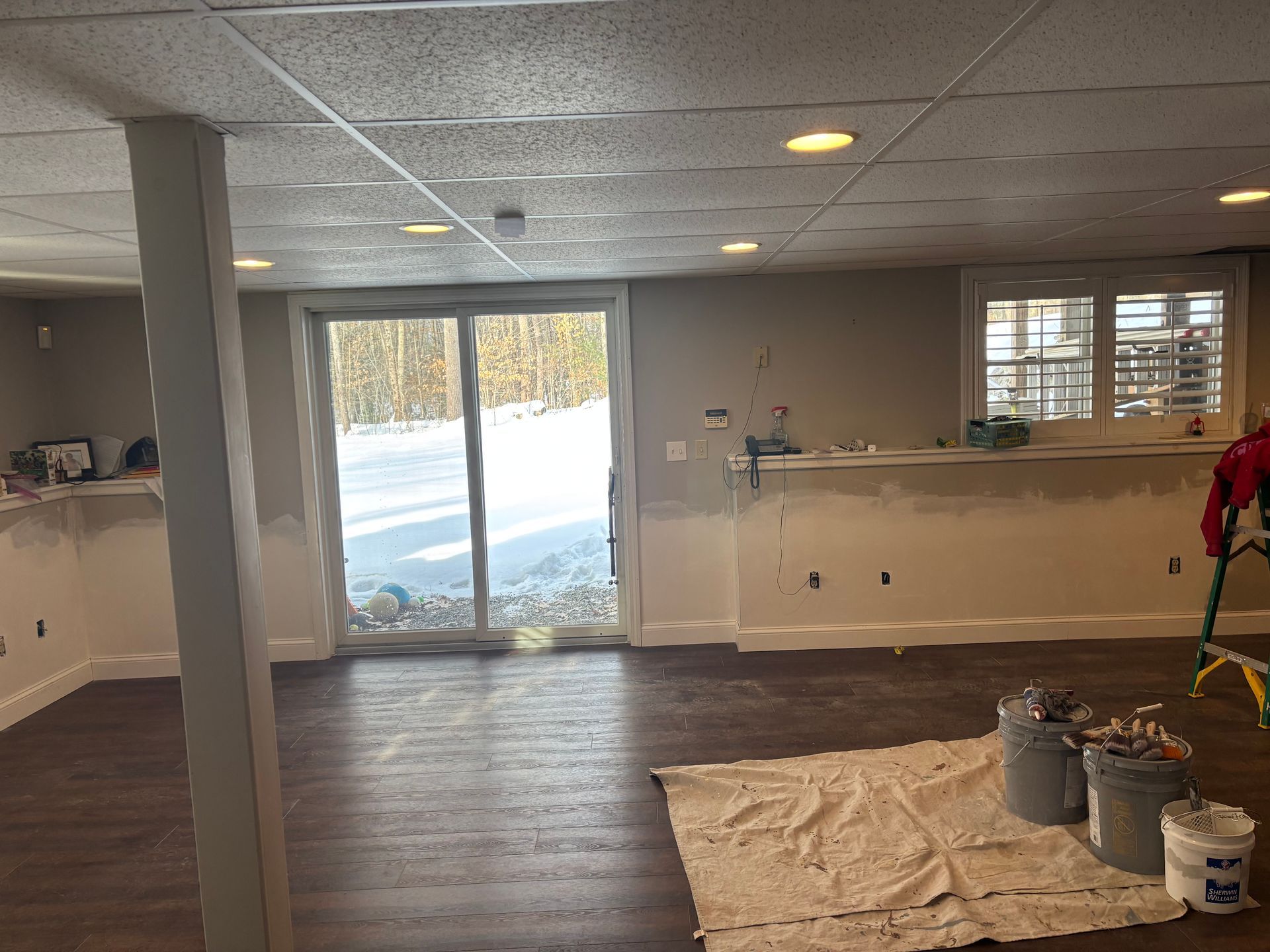 A basement interior with gray walls, dark wood-look flooring, a drop ceiling, sliding glass doors, and painting supplies.