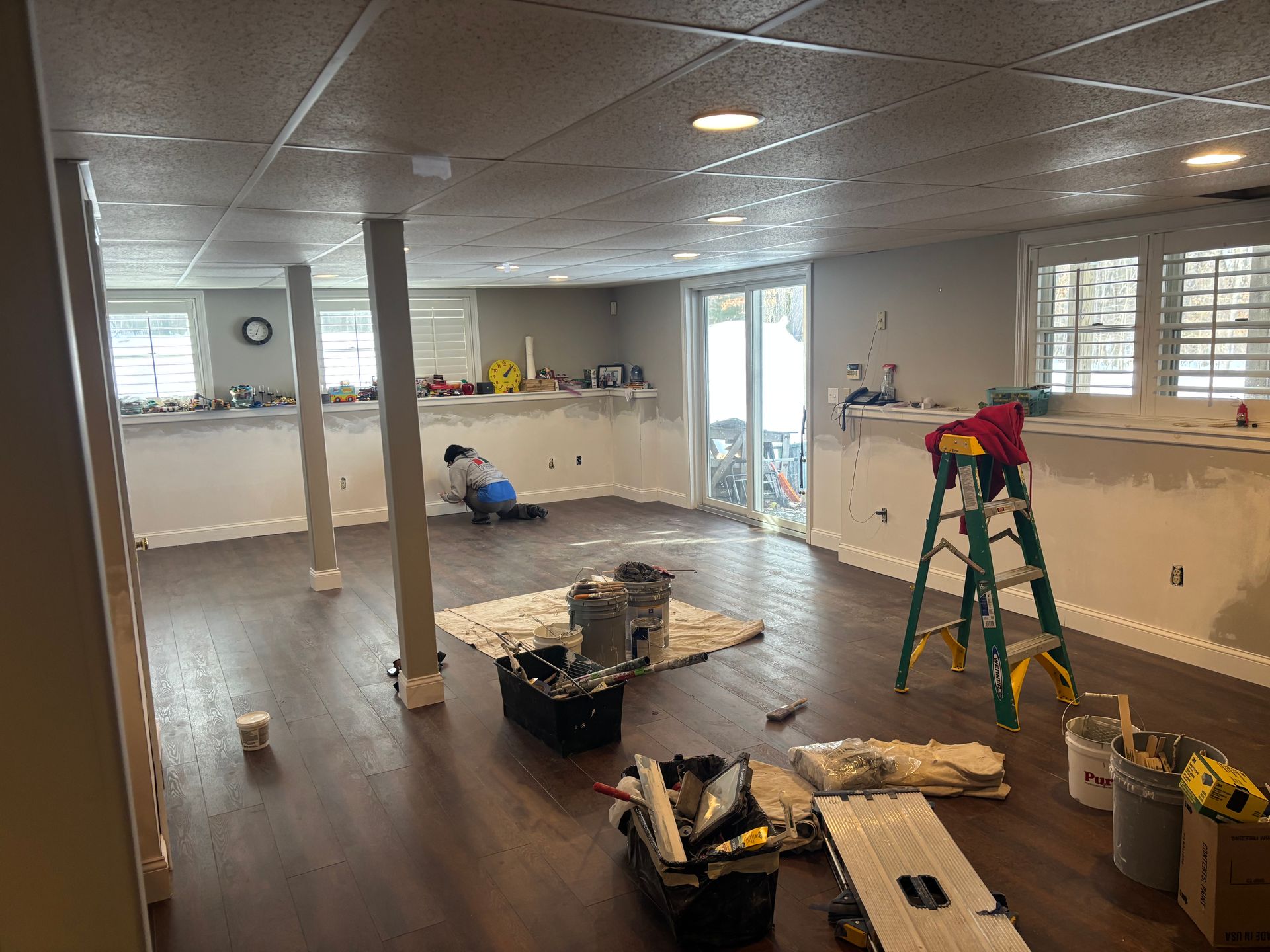 A person works on a basement renovation project featuring gray walls, dark wood-style floors, and a ladder.