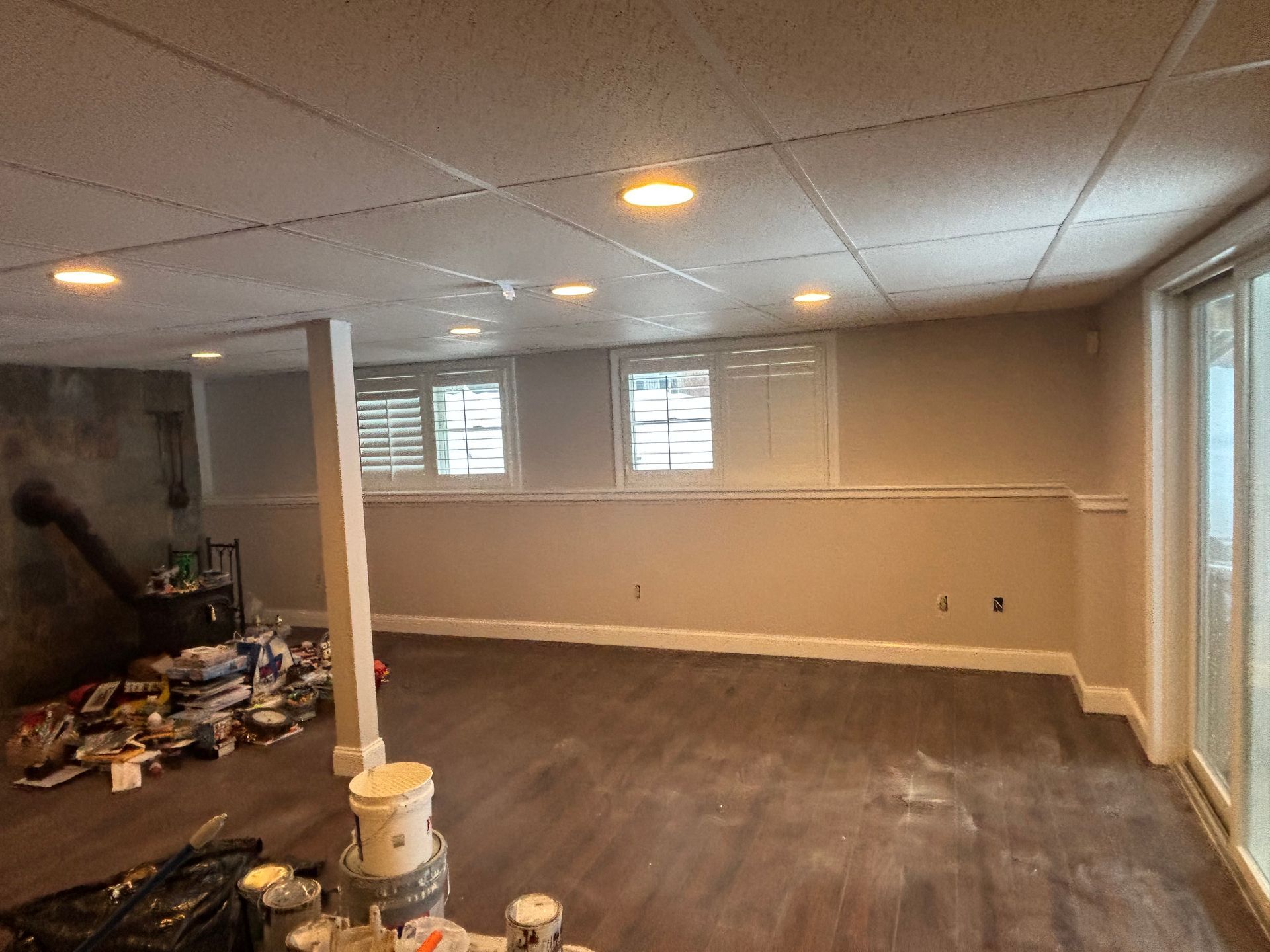 A basement interior with wood-look flooring, gray walls, recessed lighting, white window shutters, and a sliding door.