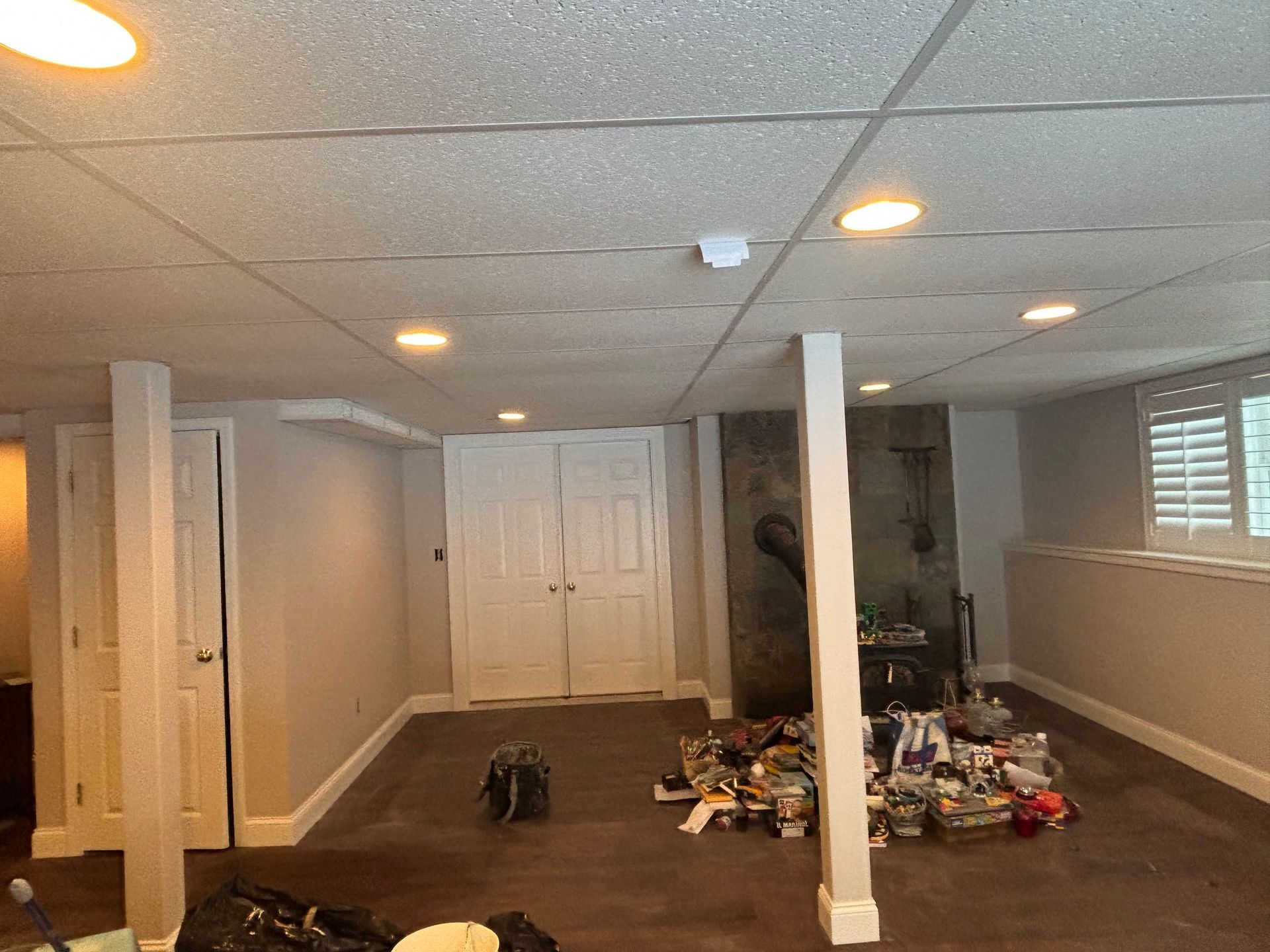 A basement with grey walls, dark flooring, white support columns, a drop ceiling with recessed lights, and some clutter.