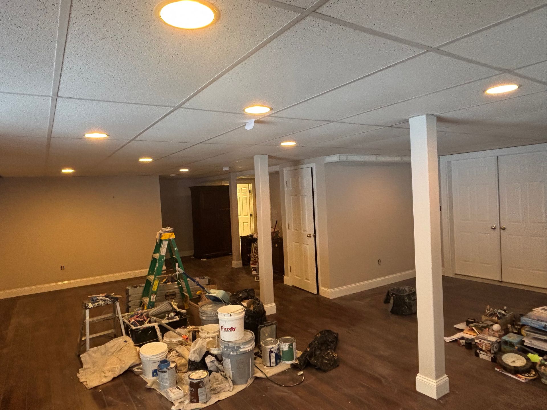 A basement renovation in progress with a ladder, painting supplies, grey flooring, white support posts, and recessed lights.
