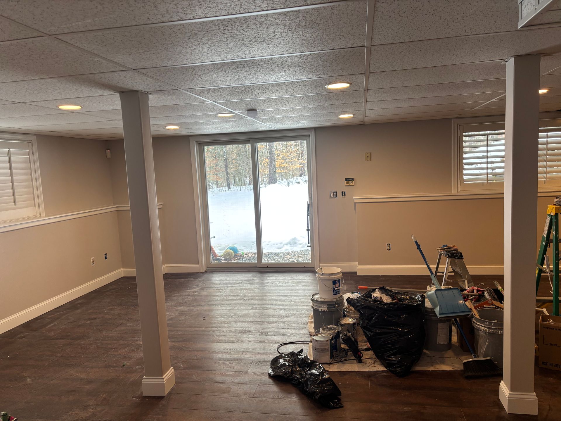 A basement room with dark wood flooring, light beige walls, two structural pillars, and a sliding glass door.