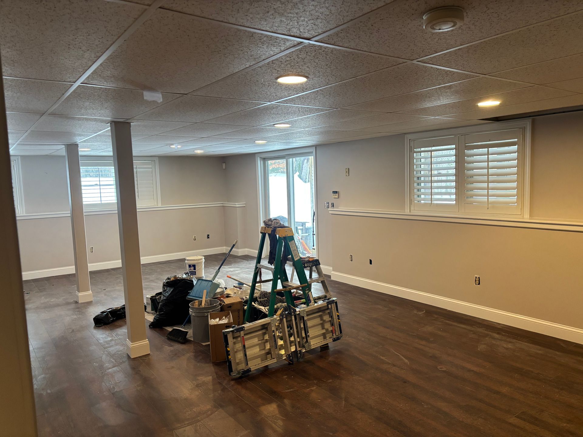 A basement under renovation with a stepladder, tools, and construction supplies in the center on dark wood-look flooring.