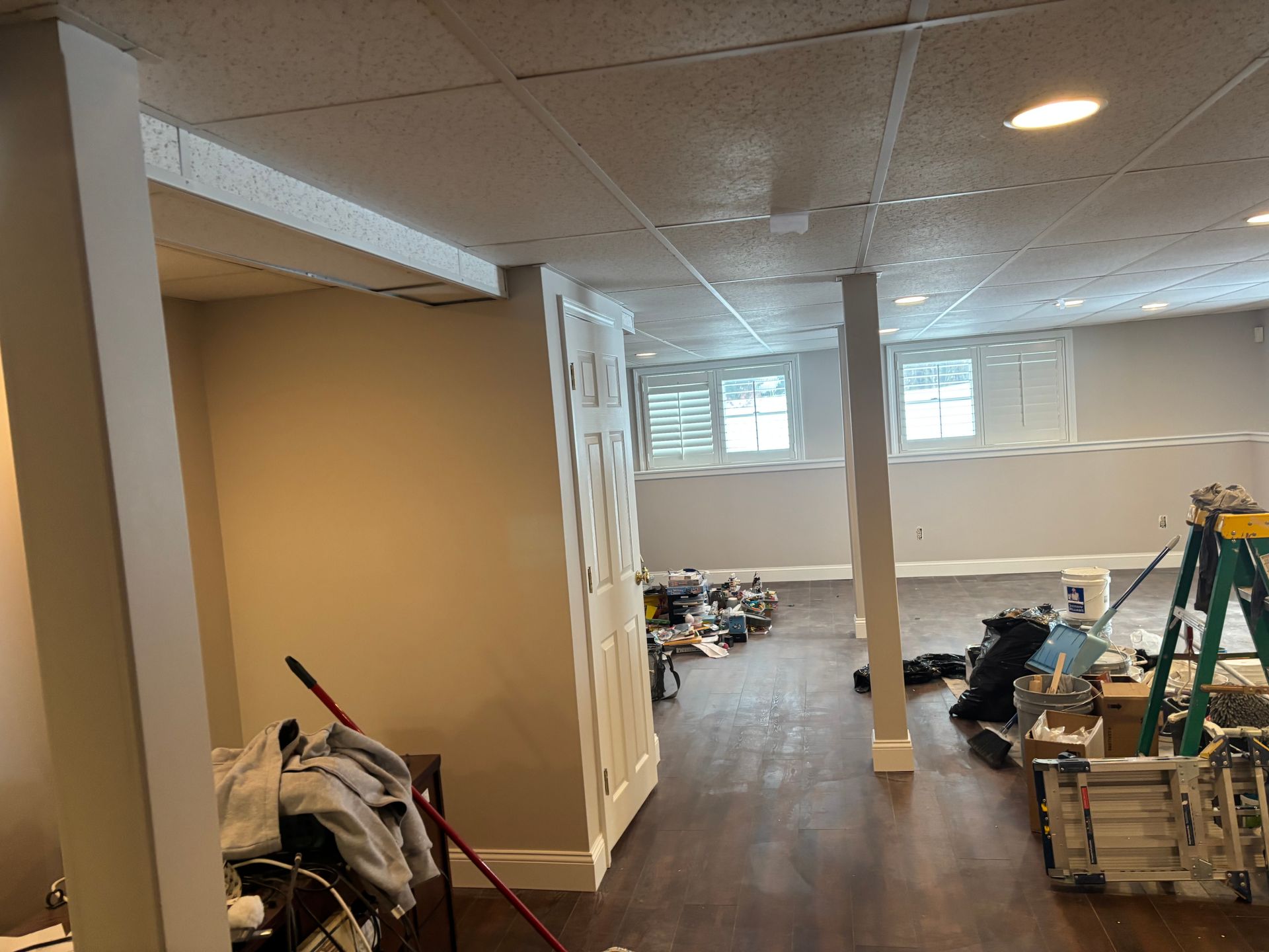 An unfinished basement with wood-look flooring, support columns, a ceiling grid, and construction tools and supplies.