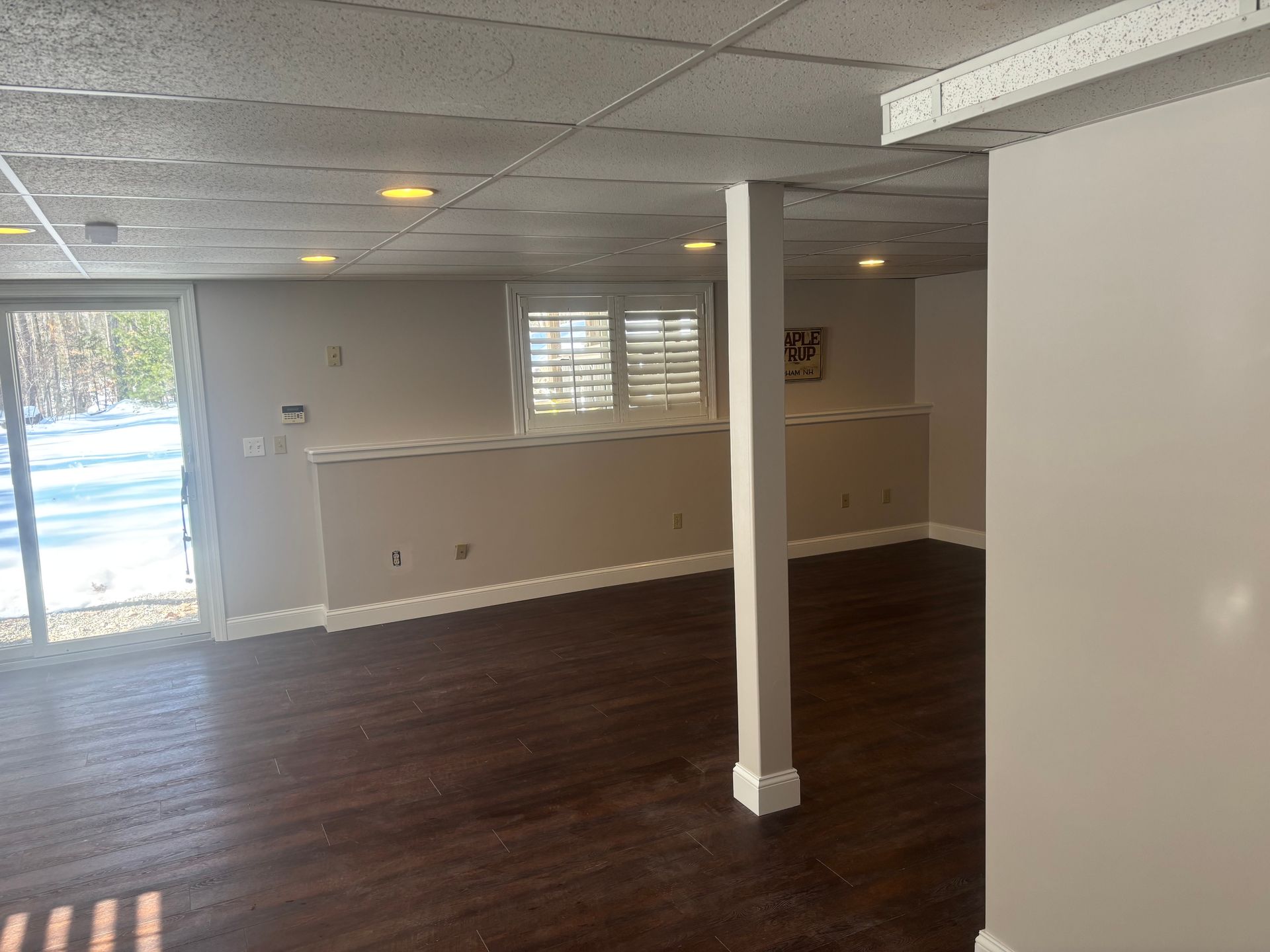 A basement interior with dark wood flooring, beige walls, a support column, recessed lighting, and a glass sliding door.