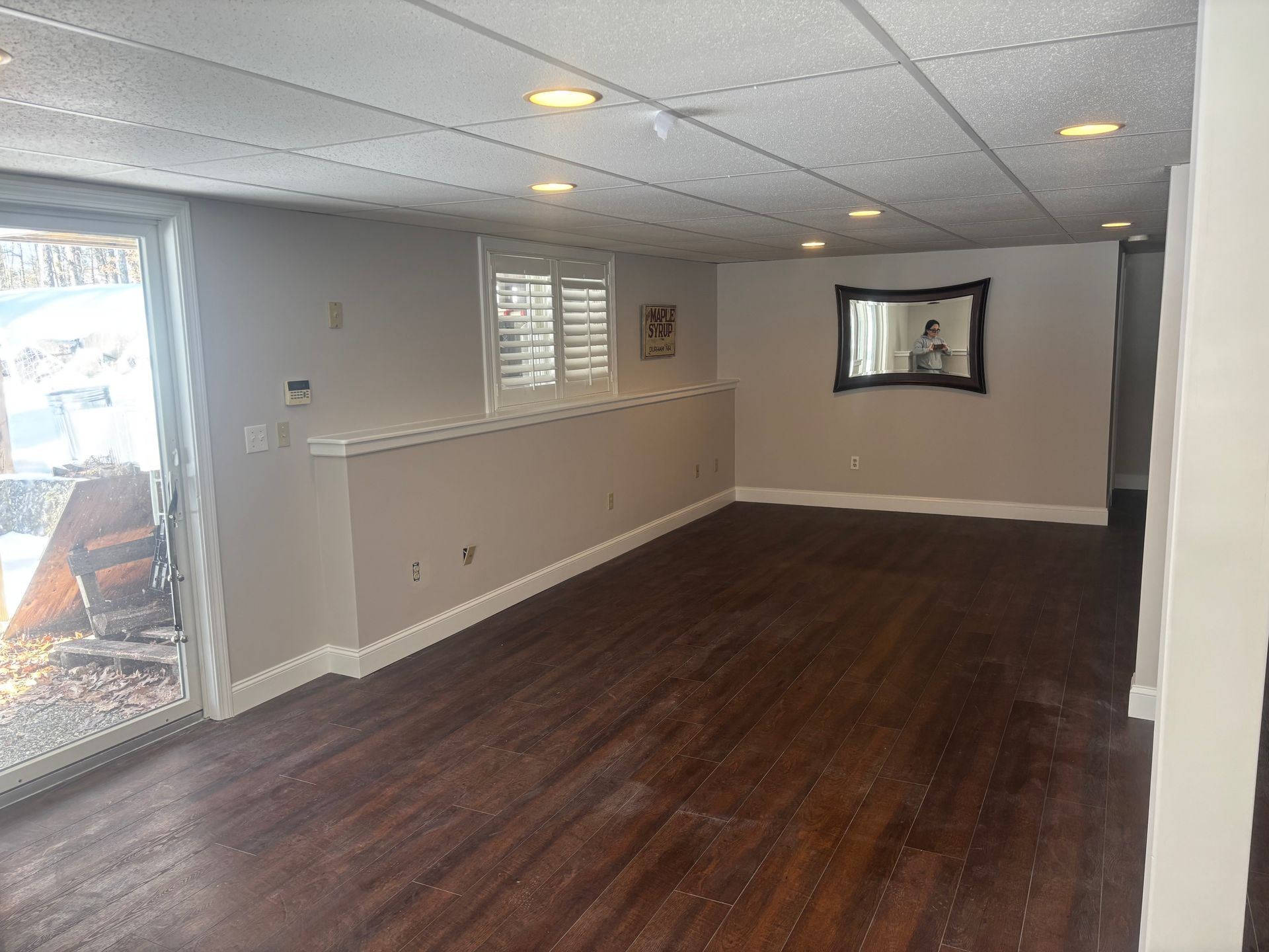 A finished basement with dark hardwood flooring, neutral walls, a drop ceiling, a sliding glass door, and a wall mirror.