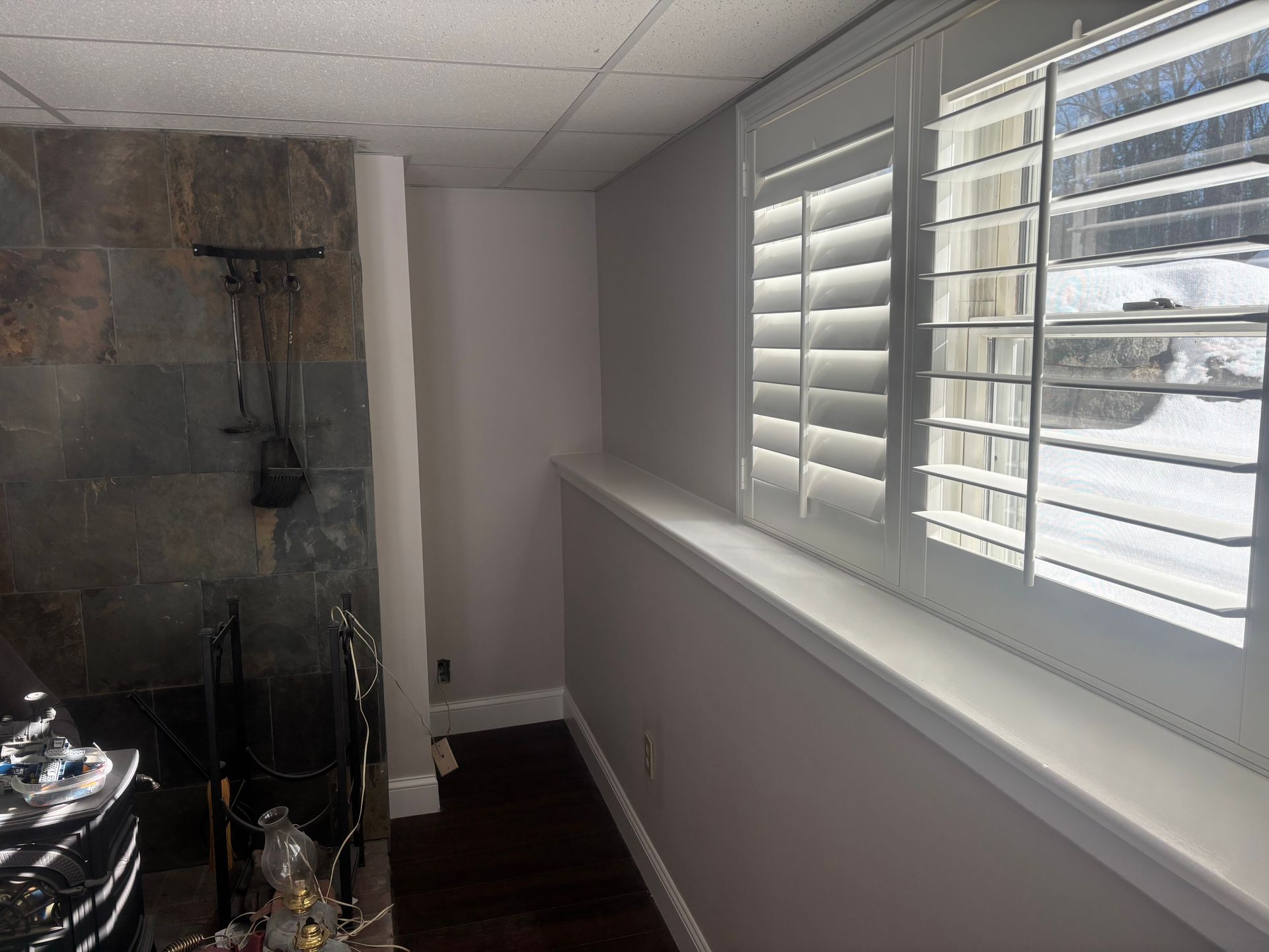 A room corner with a stone fireplace, gray walls, and a window with white shutters overlooking a snow-covered yard.