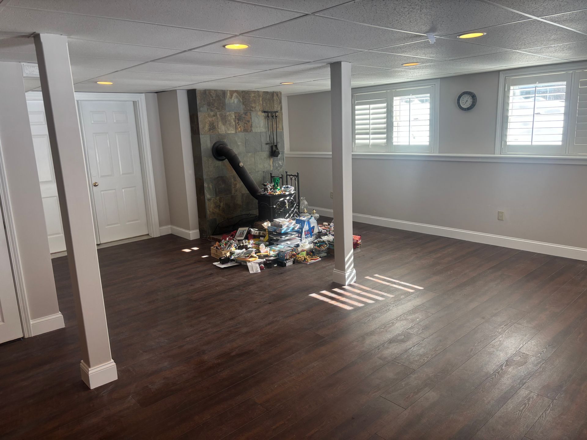 A finished basement with wood-look flooring, support pillars, a wood stove on a stone wall, and clutter on the floor.