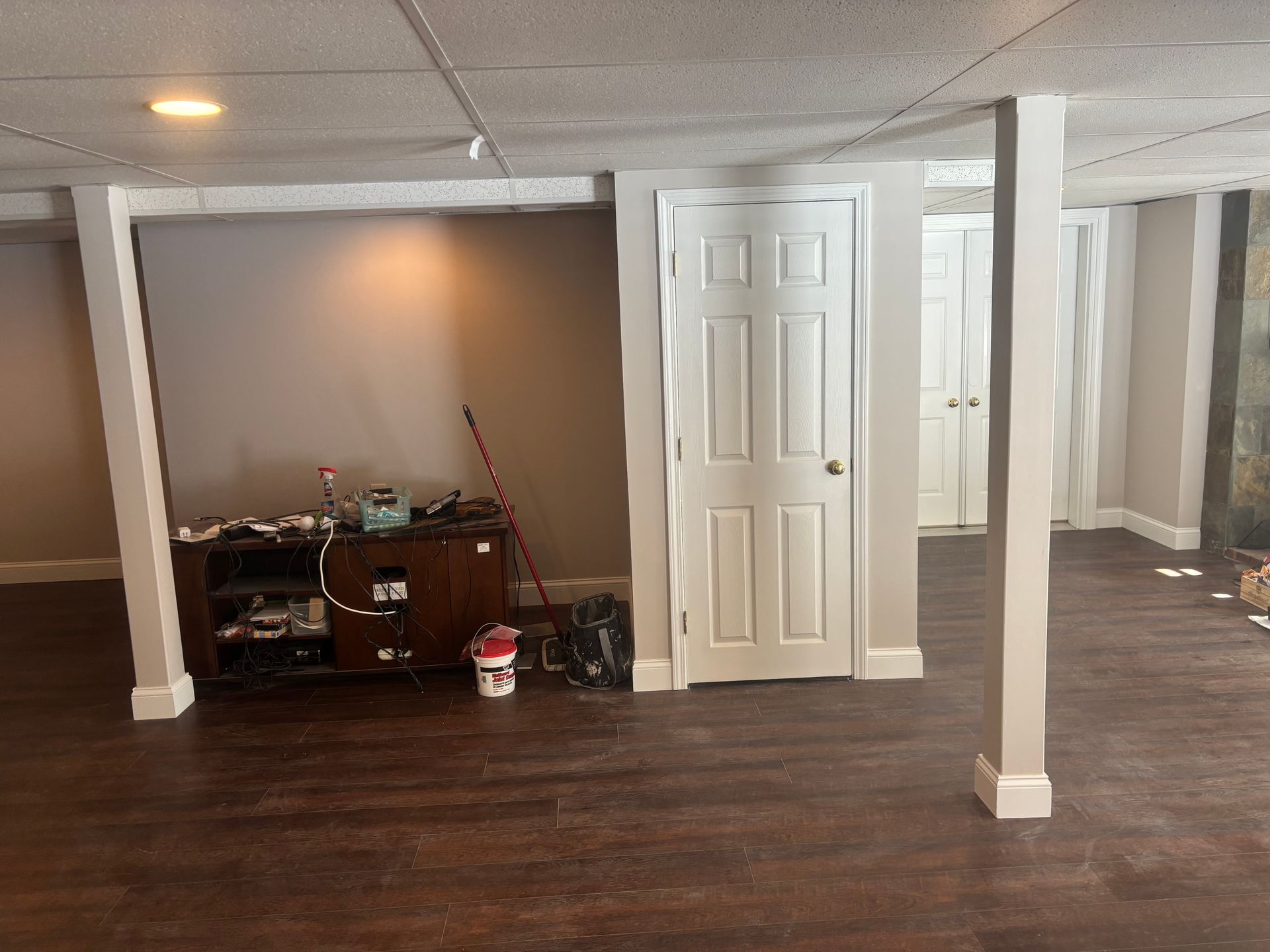 A finished basement with wood-look flooring, beige walls, two structural support pillars, and a brown media console.