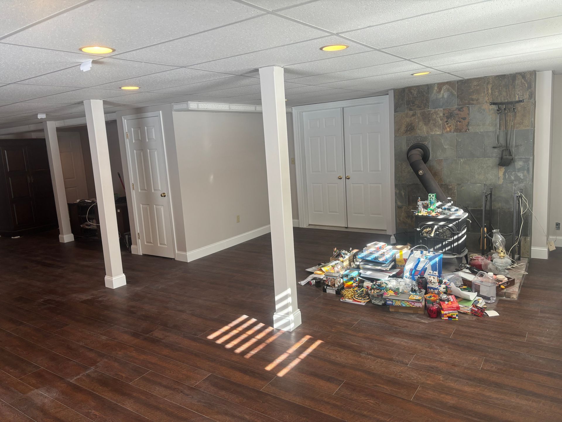 A basement room with dark wood flooring, white support pillars, ceiling panels, double doors, and a stone wood-burning stove.
