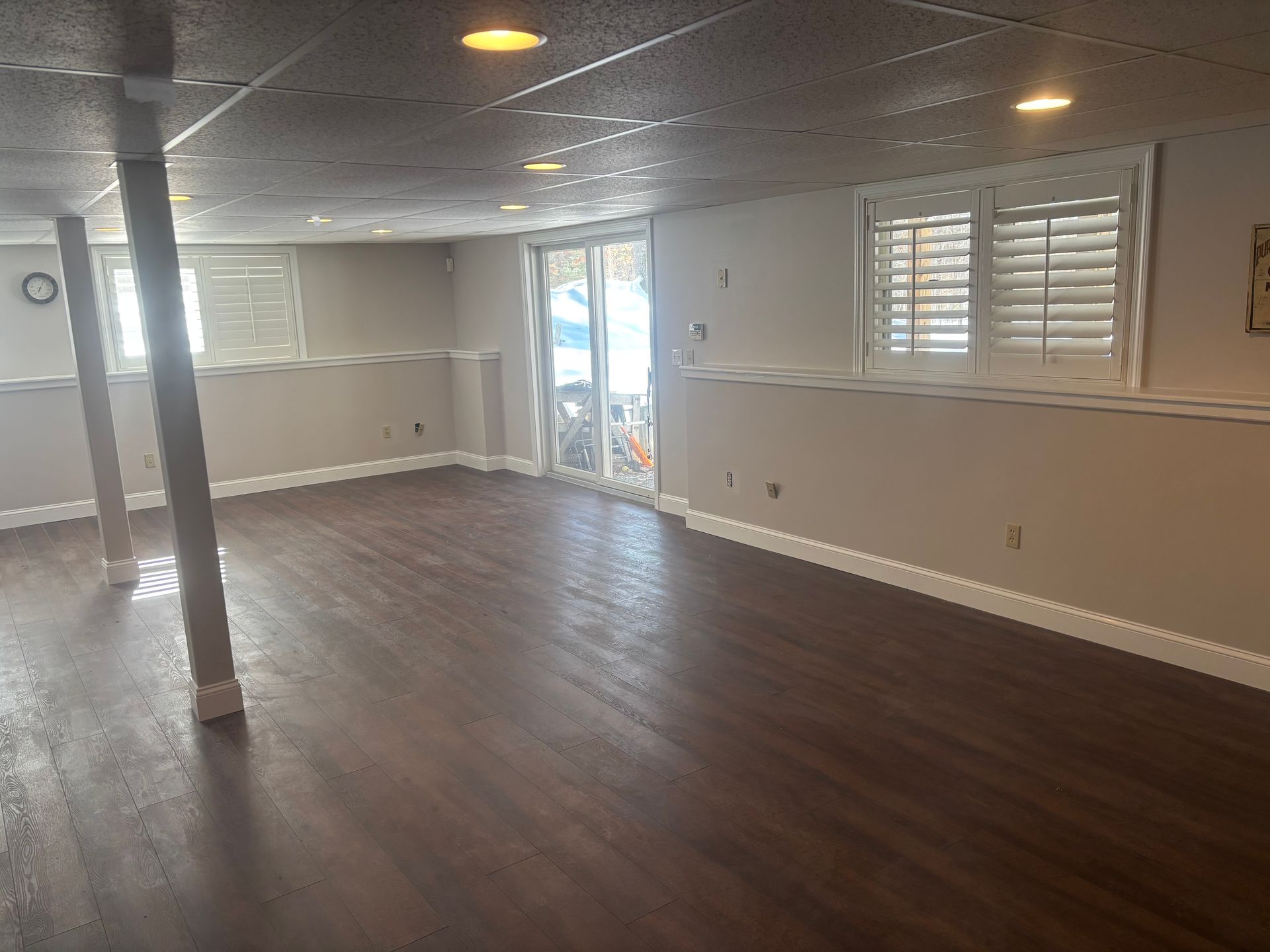 A finished basement featuring dark wood flooring, beige walls, recessed lighting, support columns, and two windows.