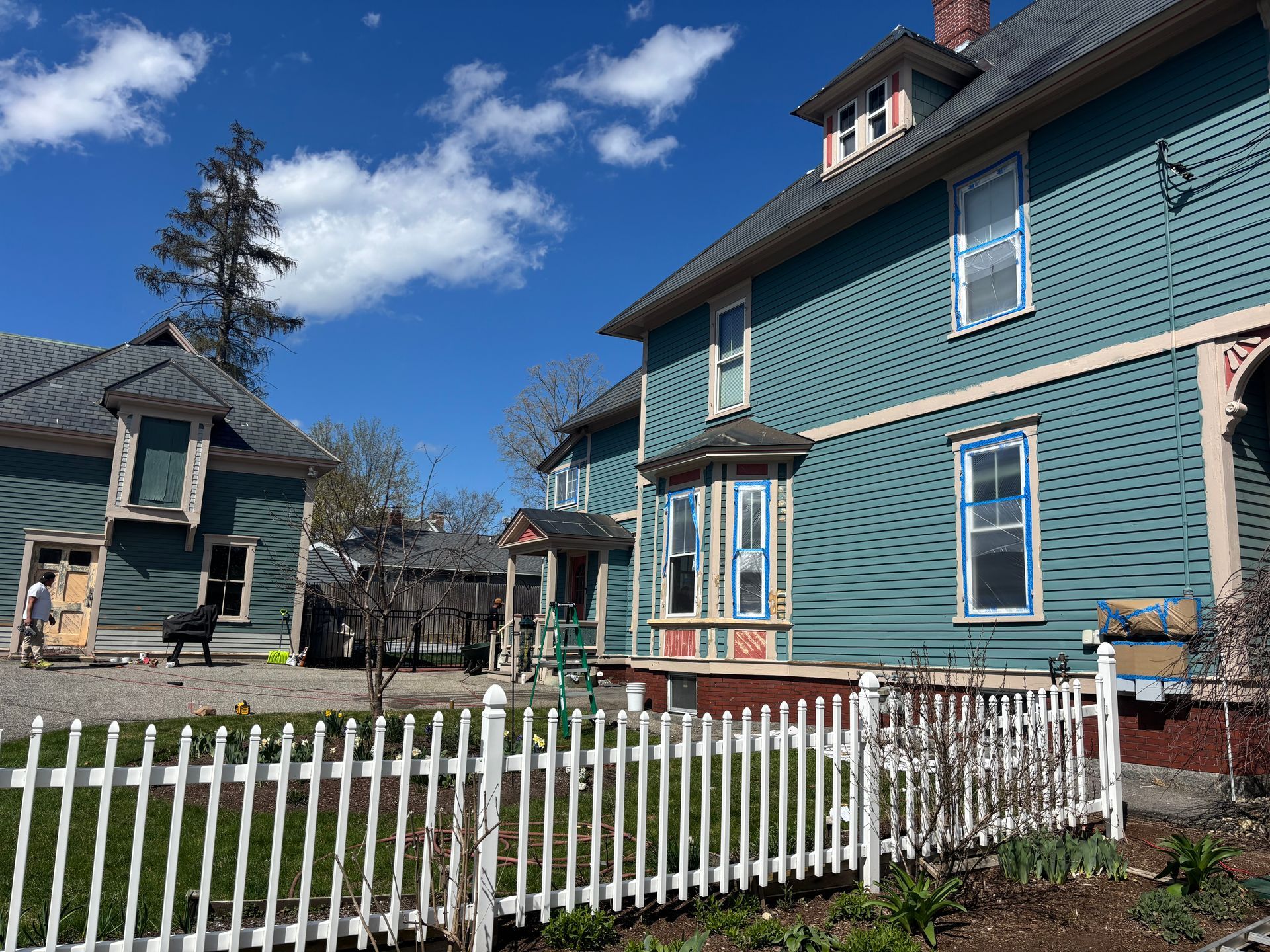 A two-story blue house under a bright blue sky, with a white picket fence in front and a smaller house to the left.