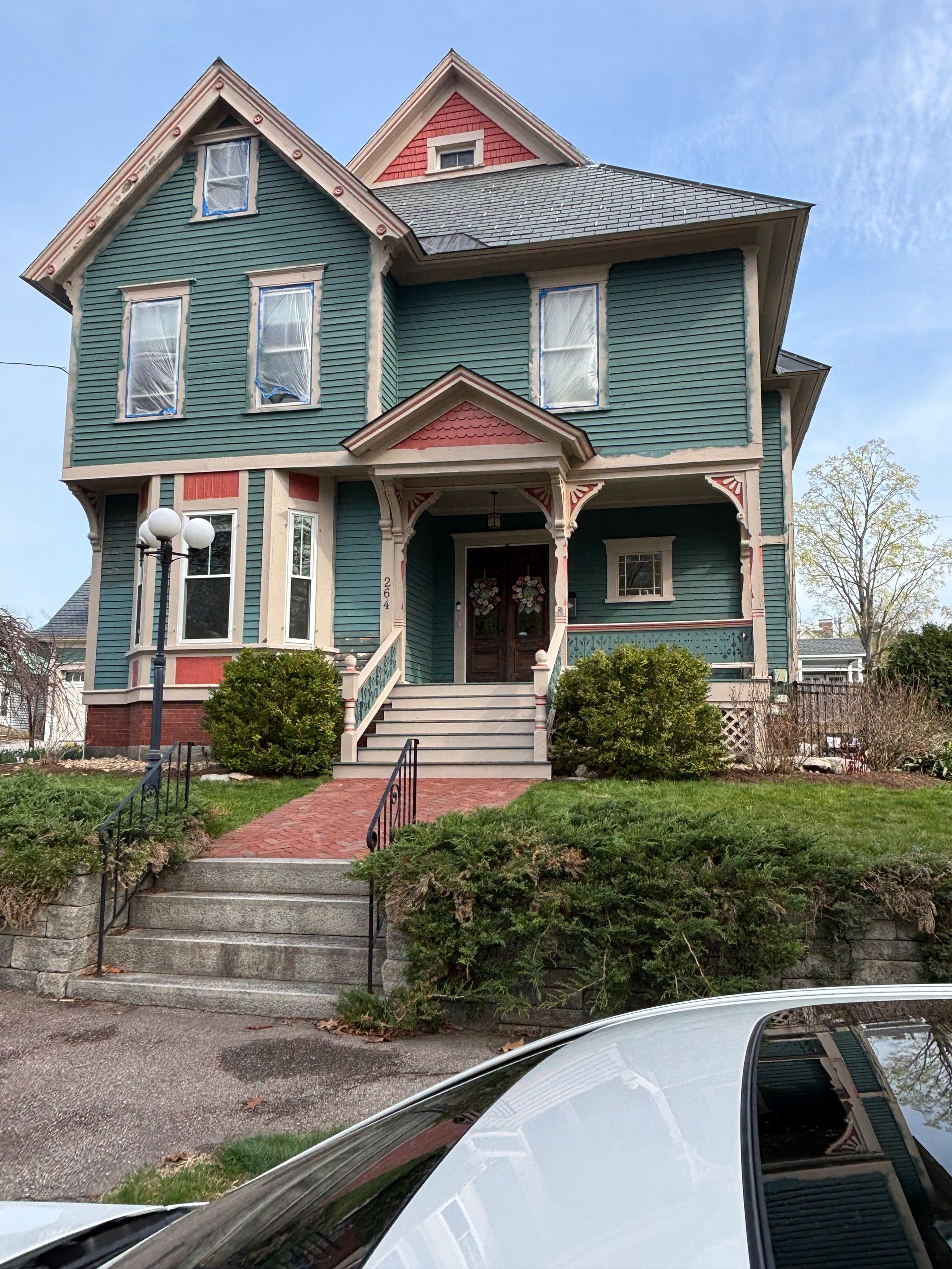 A two-story Victorian house painted teal with decorative red trim and shingled siding, viewed from a street-level path.