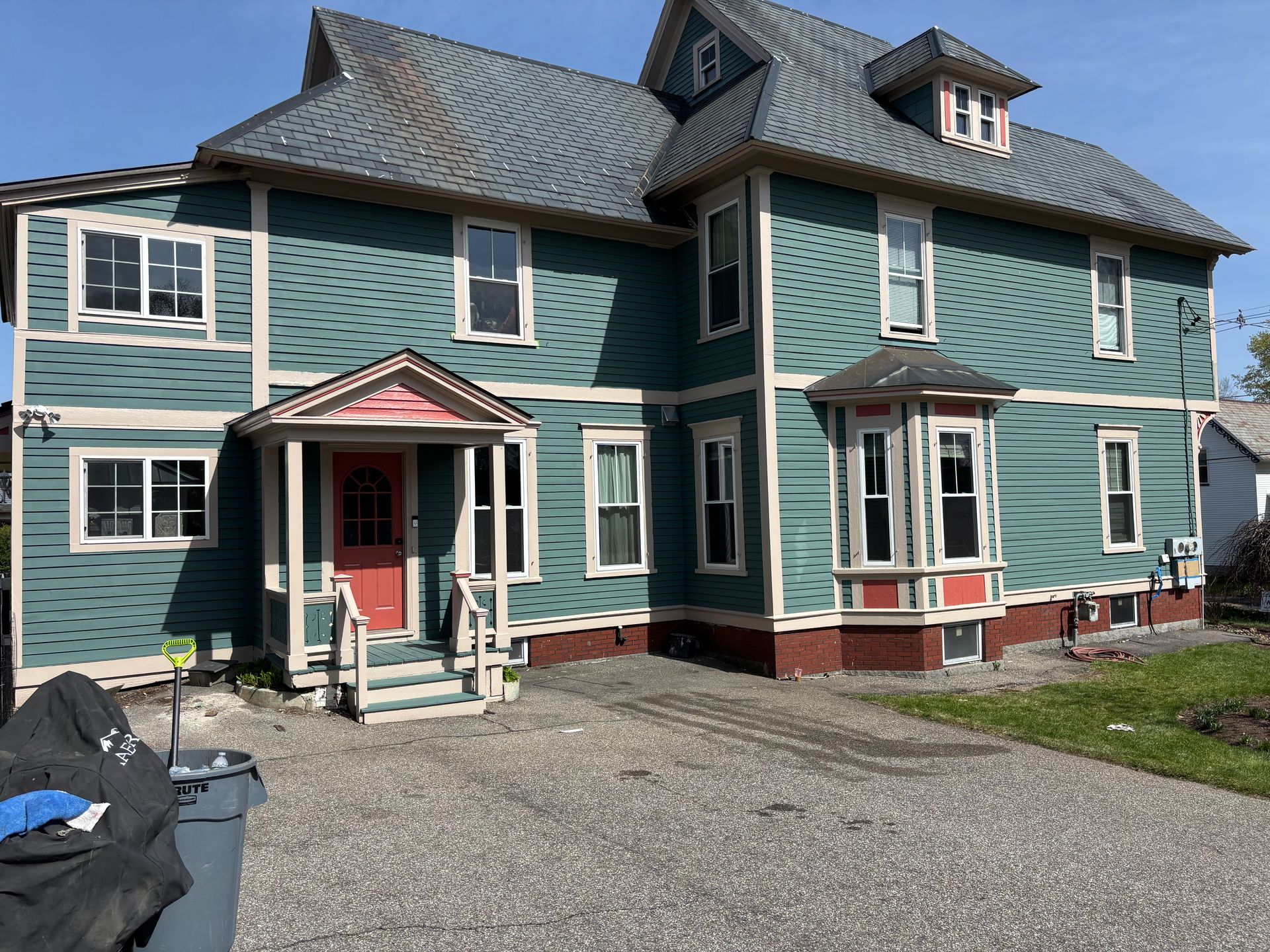 A two-story, teal-sided house with white trim, a red front door, and a gravel driveway on a sunny day.