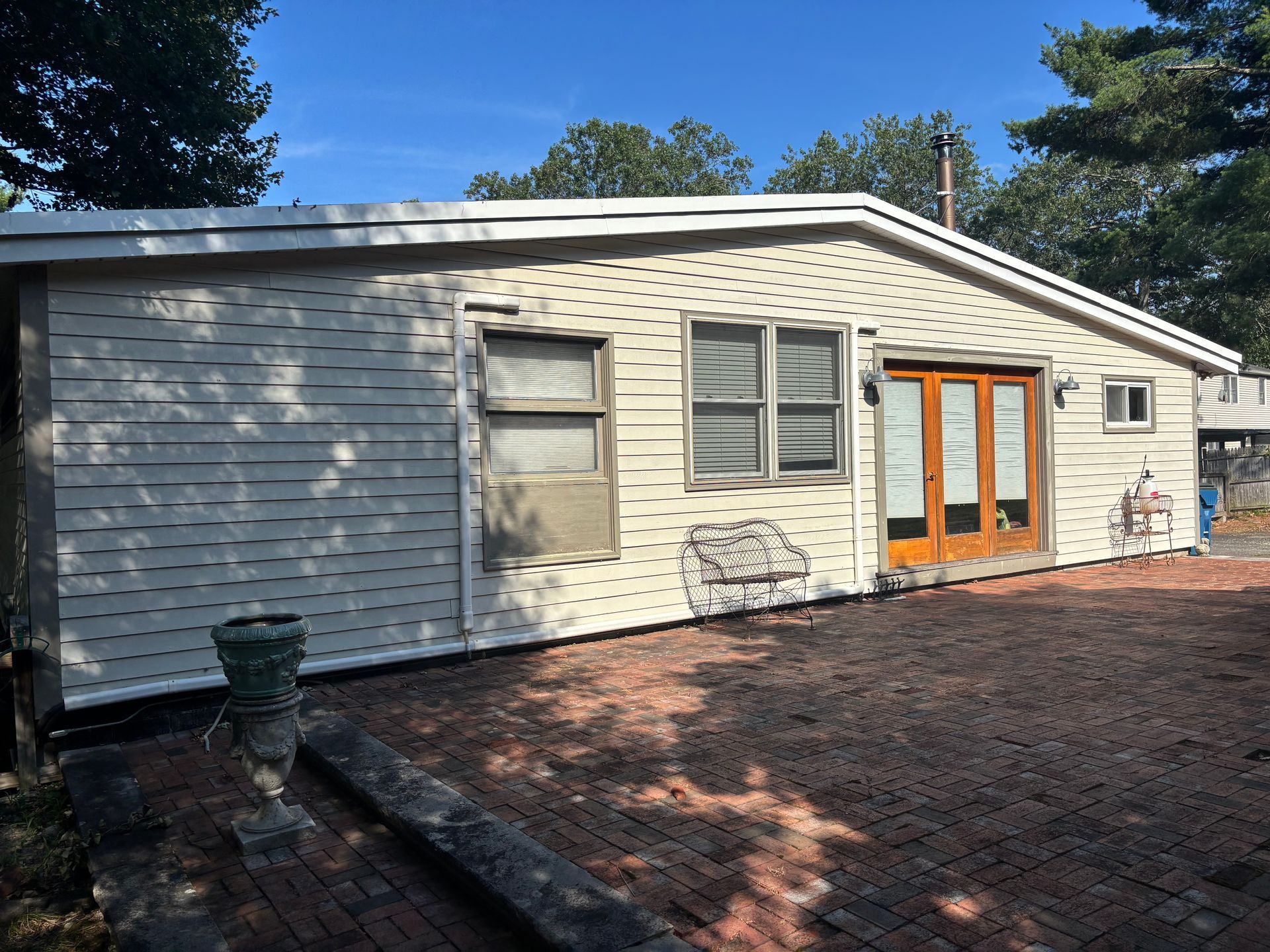 A single-story home with off-white siding, a brick patio, and wooden French doors under a clear blue sky.