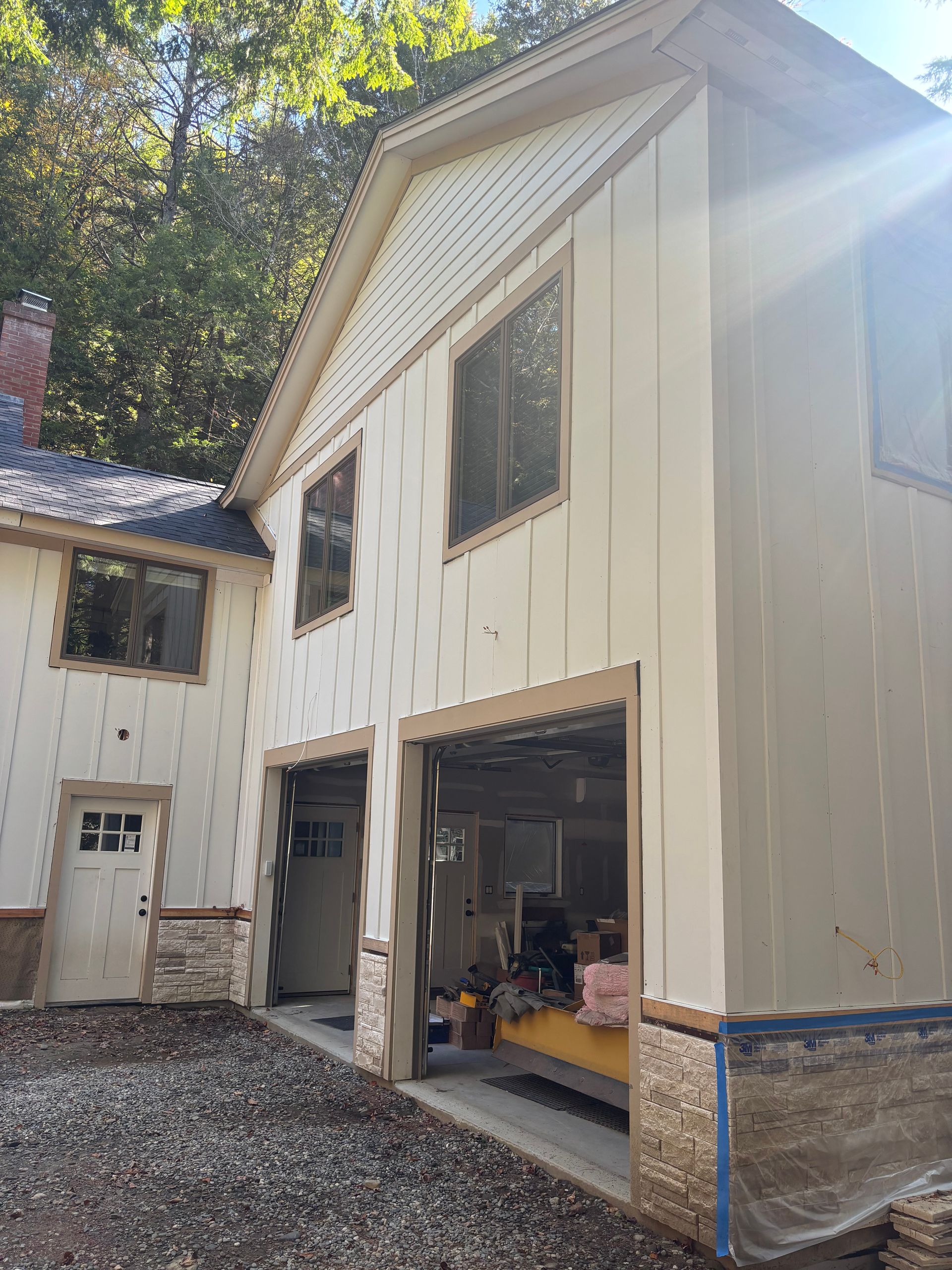 A light-colored two-story garage with vertical siding and stone accents under construction, set in a wooded area.