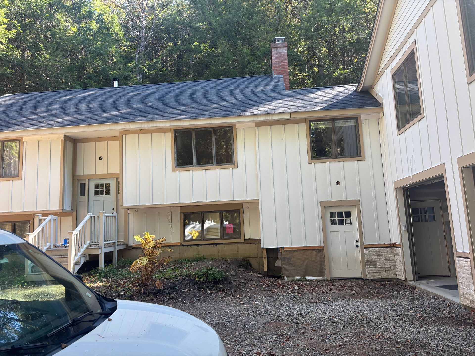 Two-story house with cream-colored vertical siding, dark roof, brick chimney, and a gravel driveway.