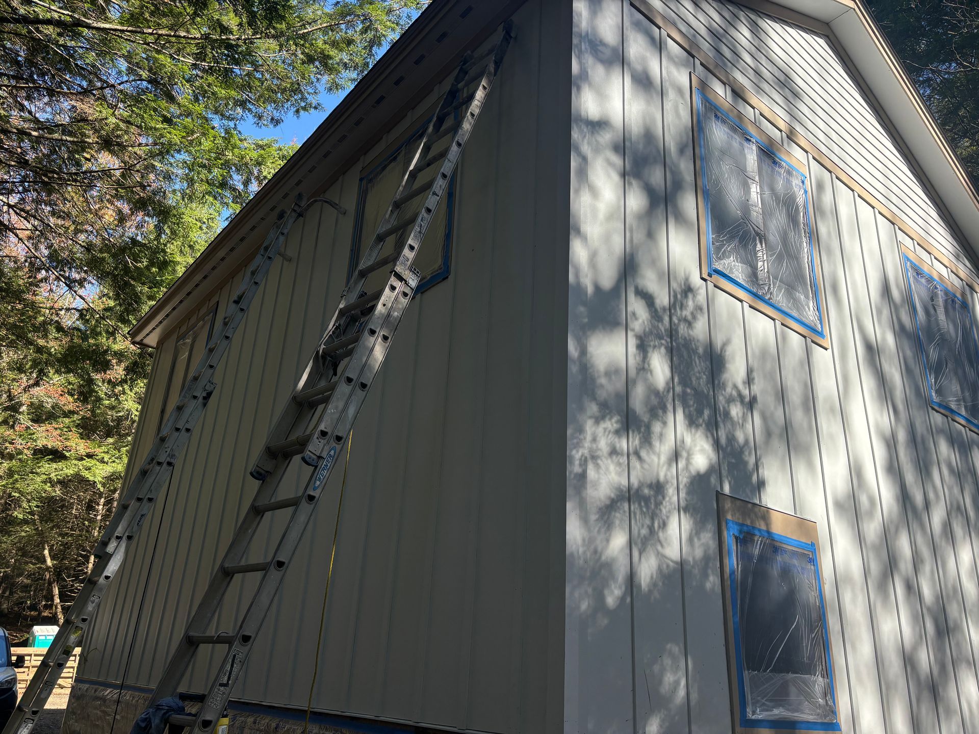 A tall, metal ladder leans against the exterior wall of a building currently being painted, with windows masked by plastic.