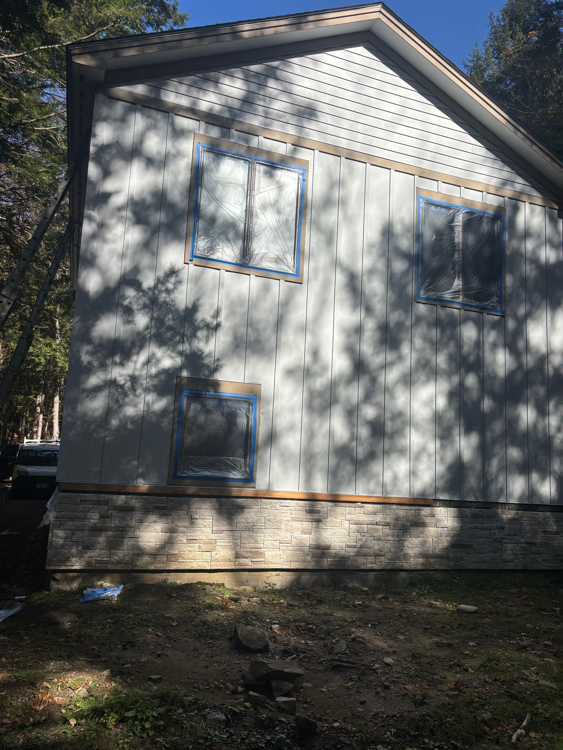 Exterior of a two-story house under renovation, featuring grey panel siding, a stone base, and new window frames.