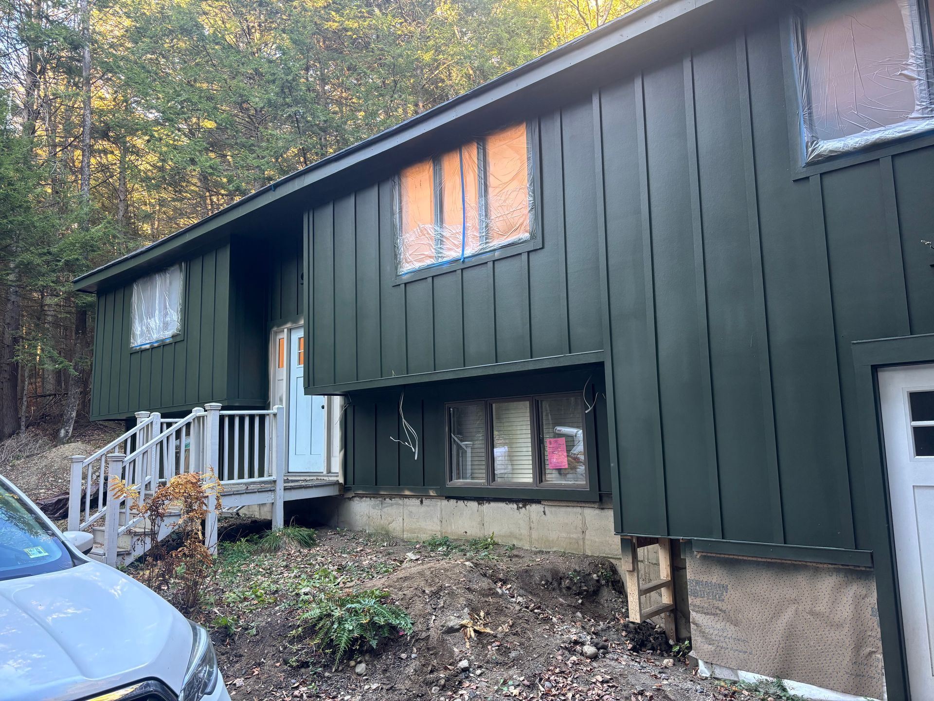 A house with dark green vertical siding, an elevated wooden porch, and windows covered with plastic sheeting.