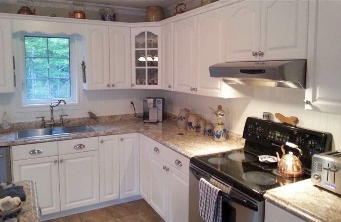 A white kitchen with wooden cabinets, granite countertops, a window over the sink, and a black stove with a vent hood.