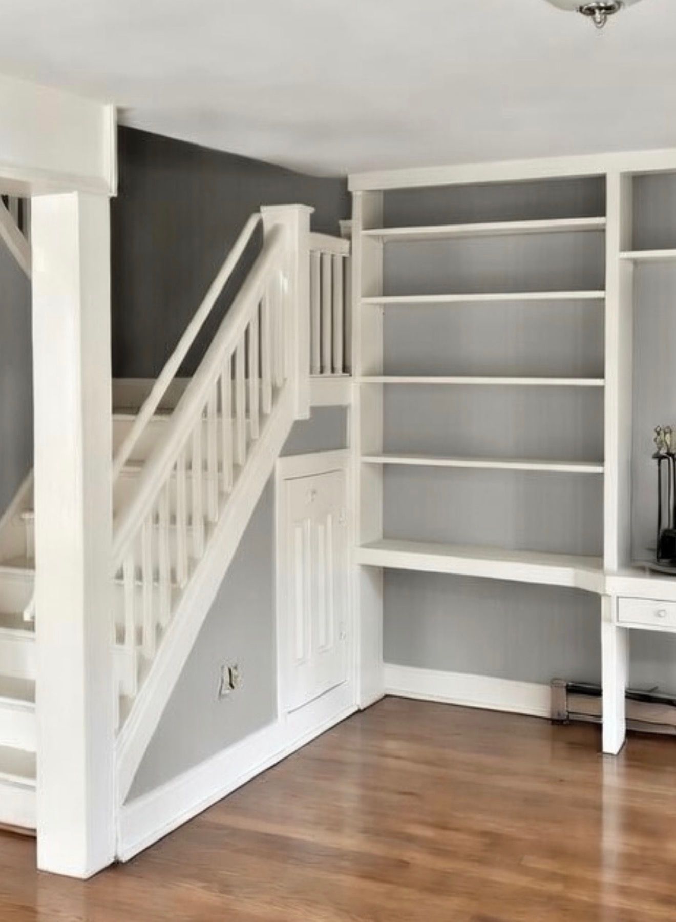 A white staircase alongside built-in white shelving and a small desk against a gray wall on hardwood floors.